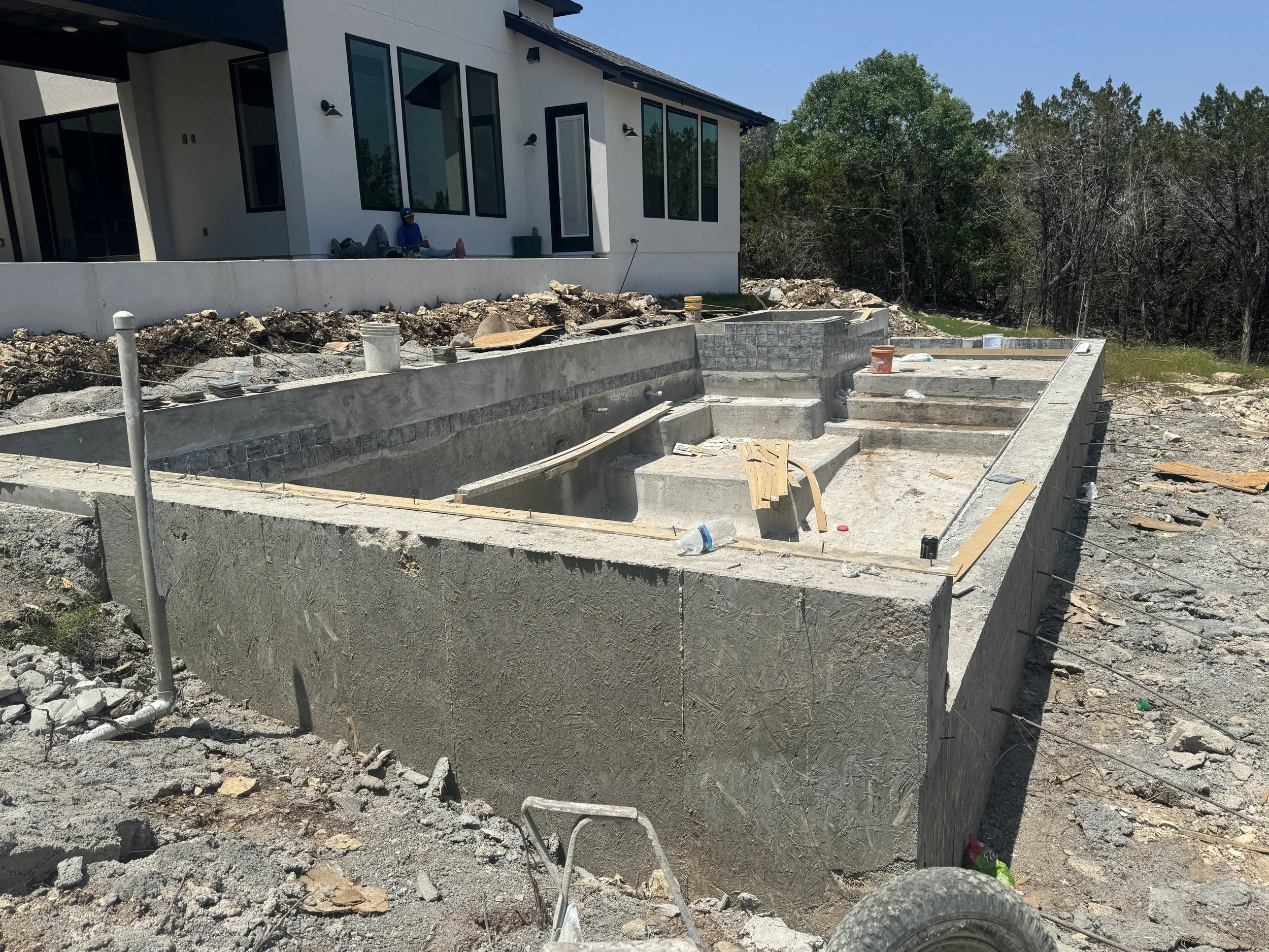 Construction site with a backyard pool in early stage, concrete structure mostly built, surrounded by construction debris, with a modern house in the background, and trees beyond.