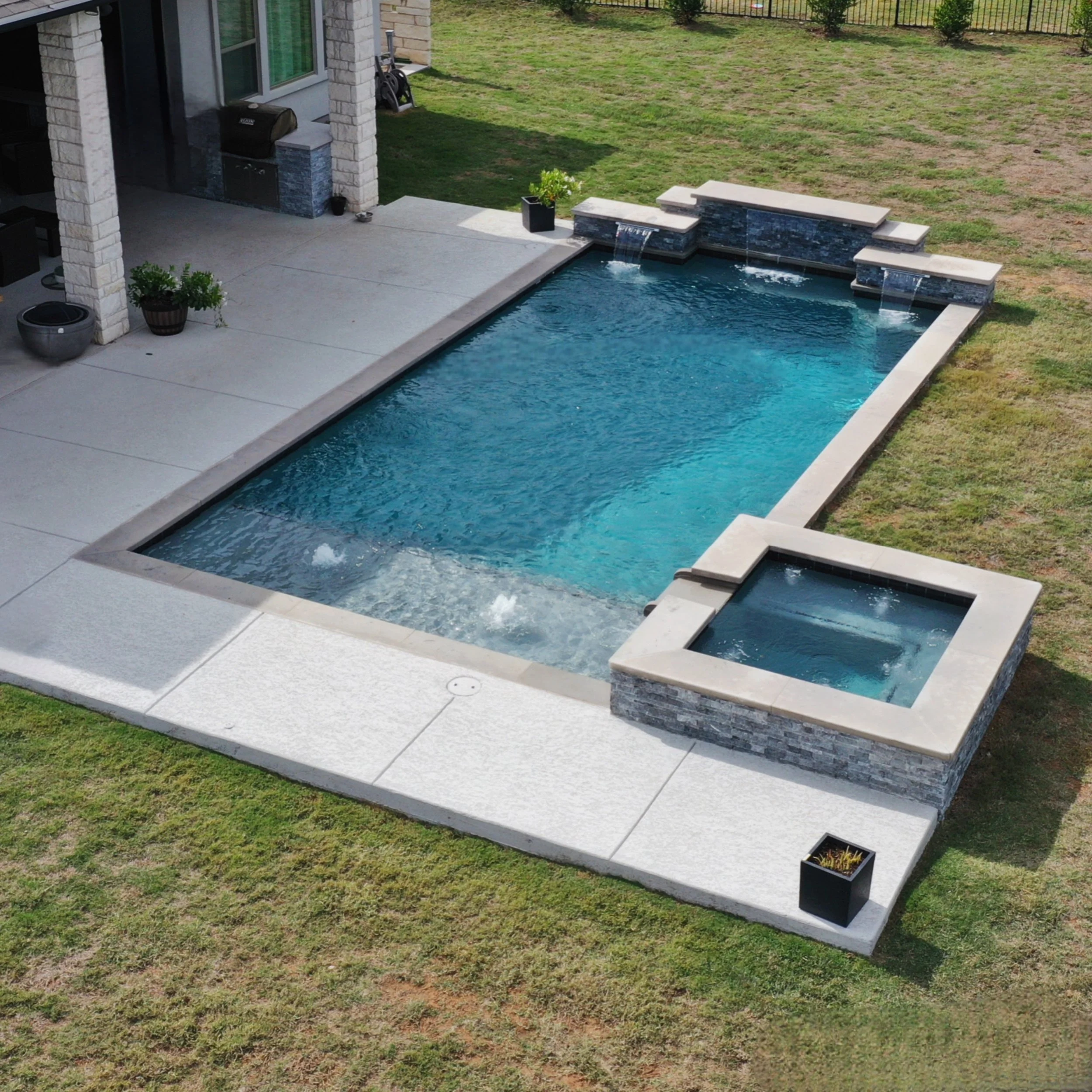 A backyard pool area with a rectangular swimming pool and a smaller attached hot tub, surrounded by a concrete deck, with a house wall and some potted plants nearby.