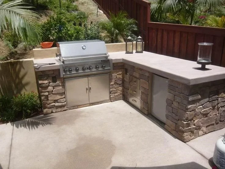 Outdoor kitchen with a gas grill, stone and concrete counter, and decorative lanterns. Lush greenery in the background.