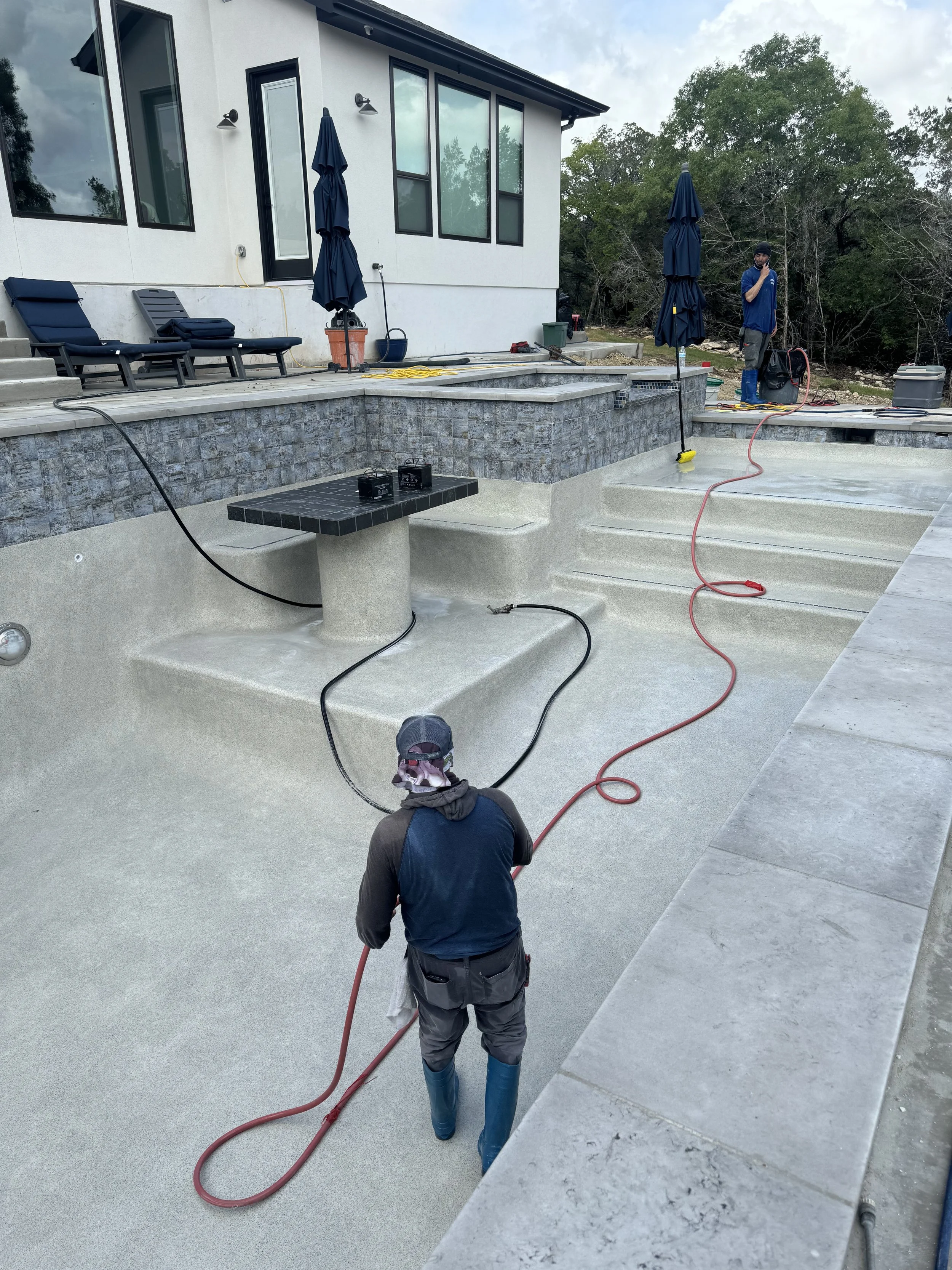 Workers applying a textured coating to a swimming pool in a backyard during daytime. The pool is empty, with steps and a small table built into the floor. The surrounding area features modern outdoor furniture, umbrellas, and a house with large windo