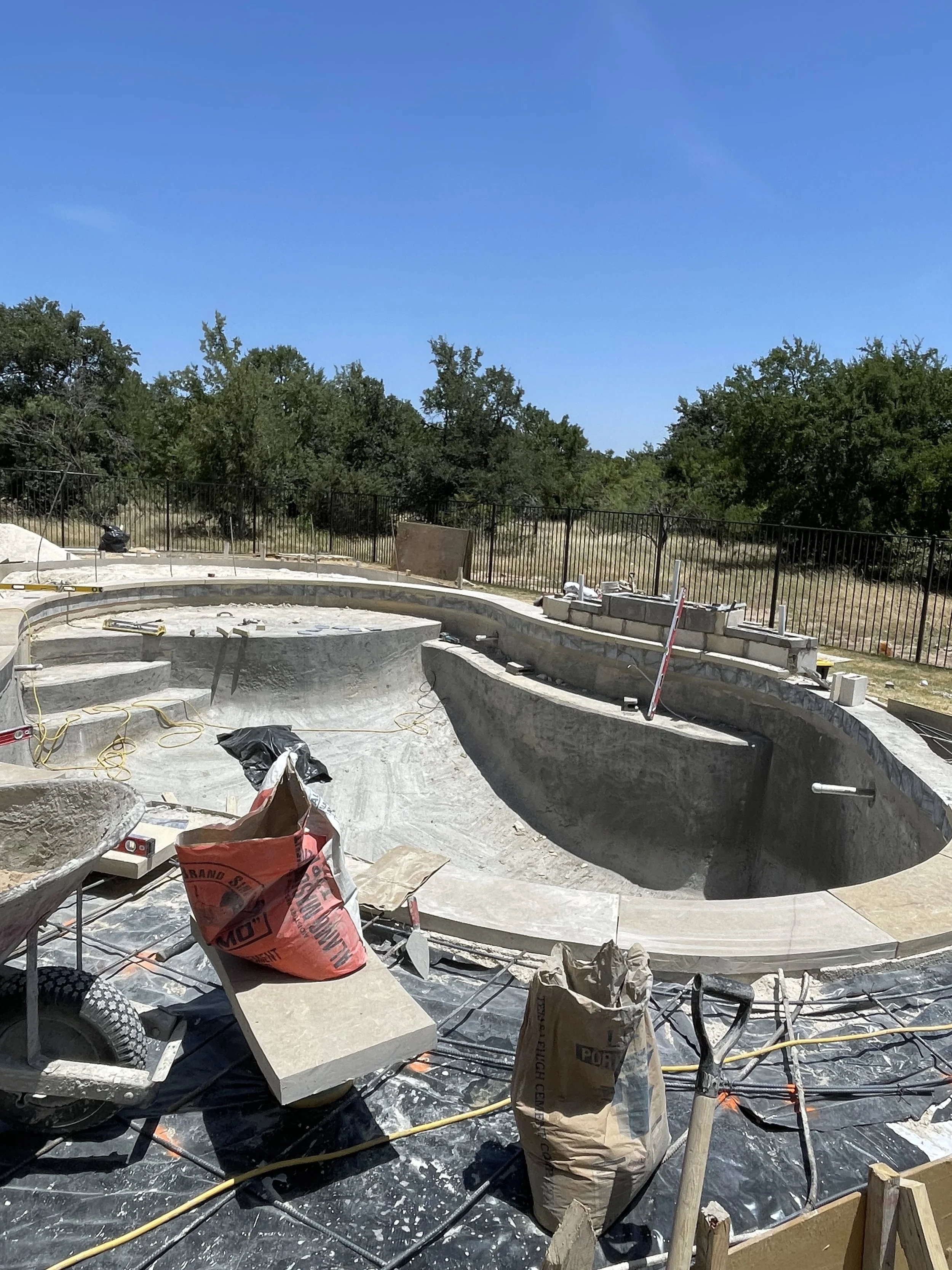 Under construction of a swimming pool with concrete shell, construction tools, bags of materials, and a black safety cover, surrounded by trees and a blue sky.