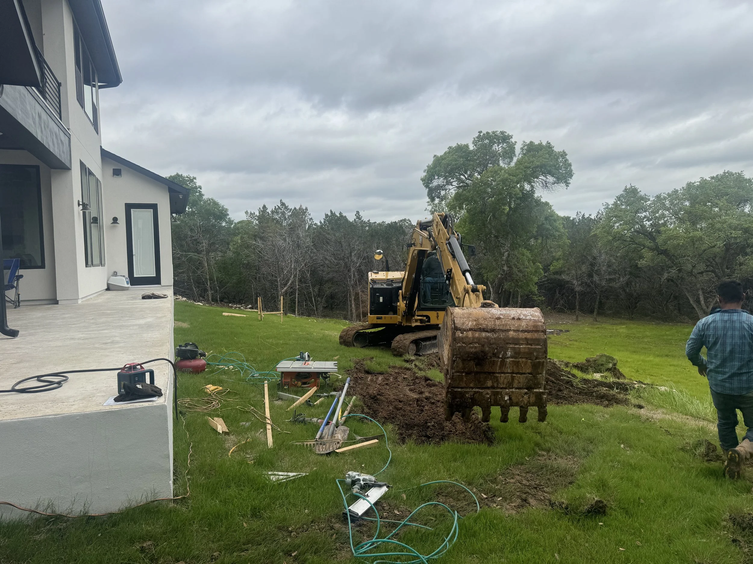 A construction site next to a house with a small excavator digging a trench on a grassy yard, with tools and equipment scattered around, and a person walking nearby under a cloudy sky.