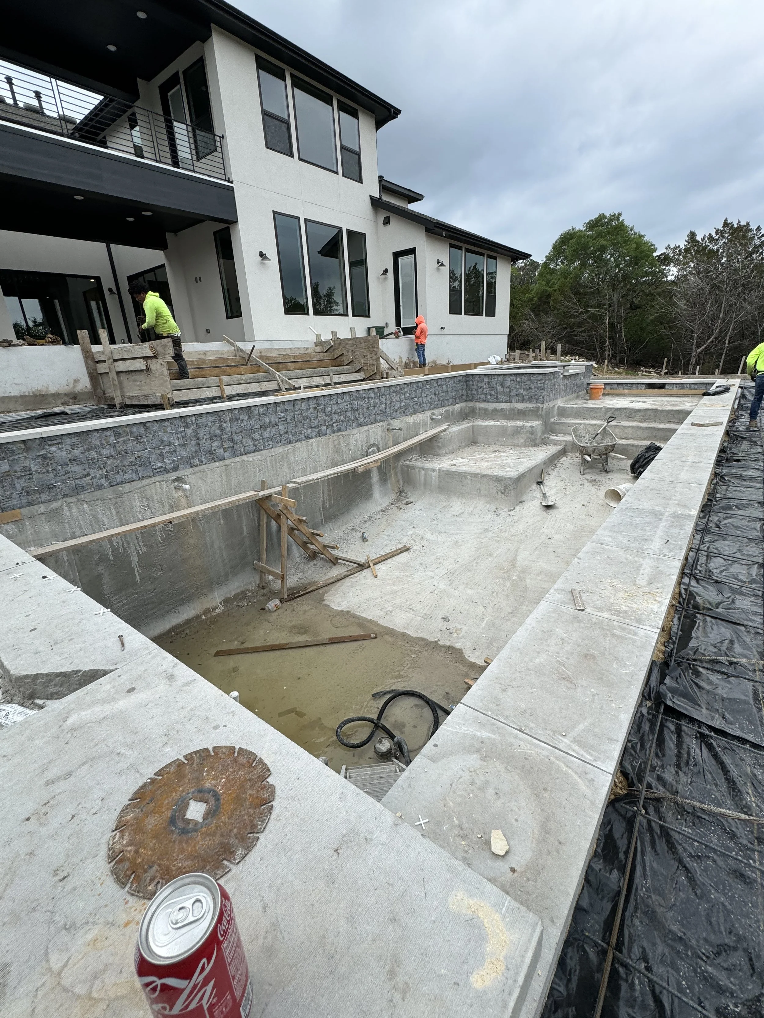 Under construction of a swimming pool in backyard of a modern house, with construction workers present, some wearing neon jackets, and construction tools and materials visible.