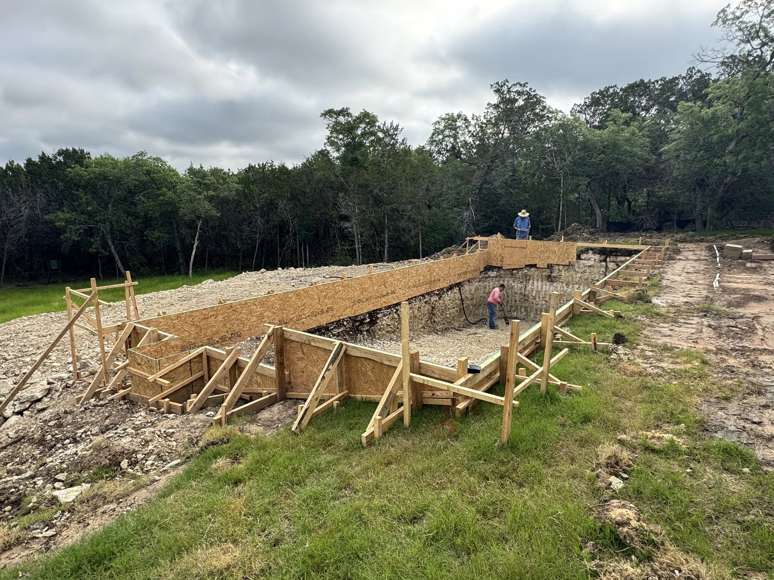 Construction workers are building a retaining wall with wooden boards and braces in a grassy area surrounded by trees, with cloudy skies overhead.