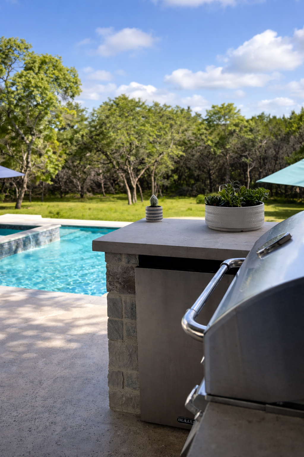 A backyard scene with a swimming pool, outdoor furniture, potted plants, and a grill, surrounded by green trees under a blue sky with clouds.