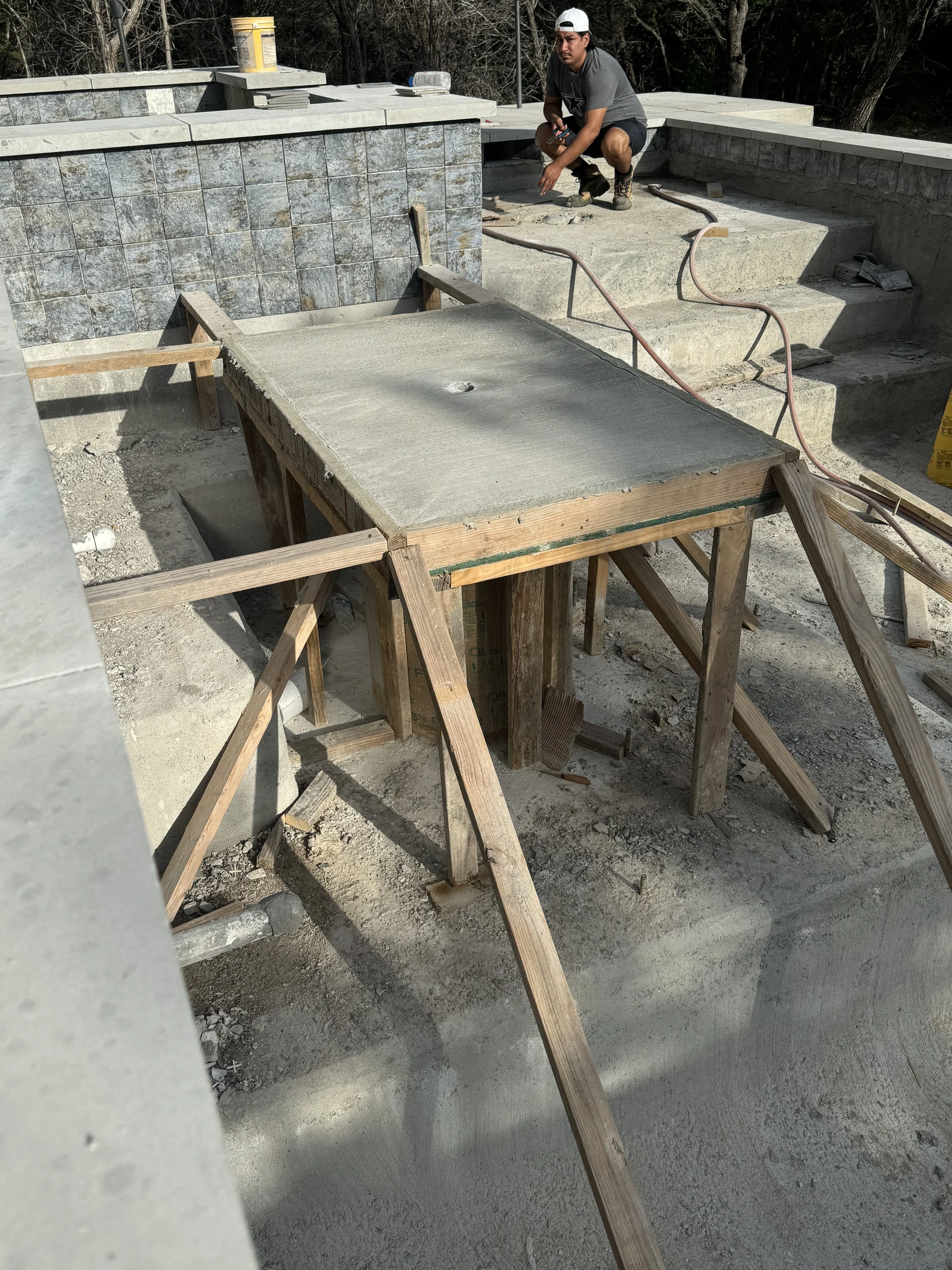 Construction site with a worker squatting near a partially built swimming pool. The worker is wearing a gray t-shirt, shorts, a white cap, and work boots. There is a freshly poured concrete slab on a wooden form and some construction equipment around