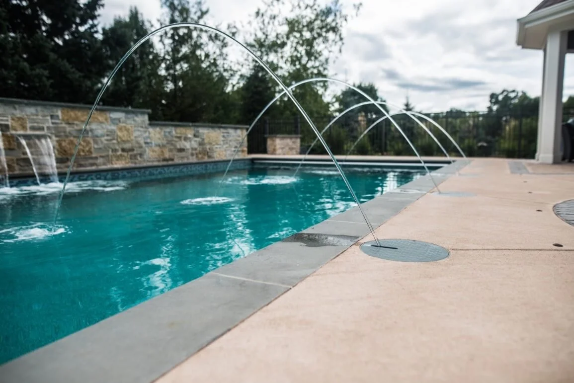 A swimming pool with four arched water jets spraying into the pool, surrounded by a stone and concrete deck, with trees and a fence in the background.
