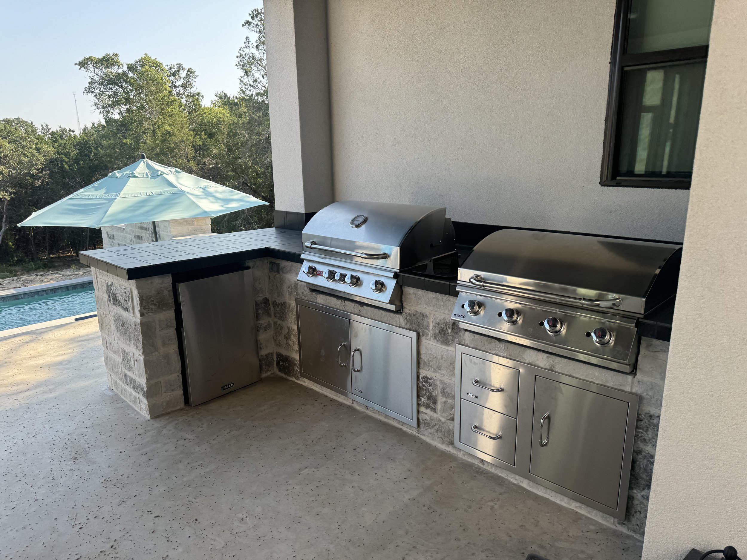Outdoor kitchen with two stainless steel gas grills, a small cabinet, and a counter with a blue umbrella nearby, overlooking a swimming pool and trees.