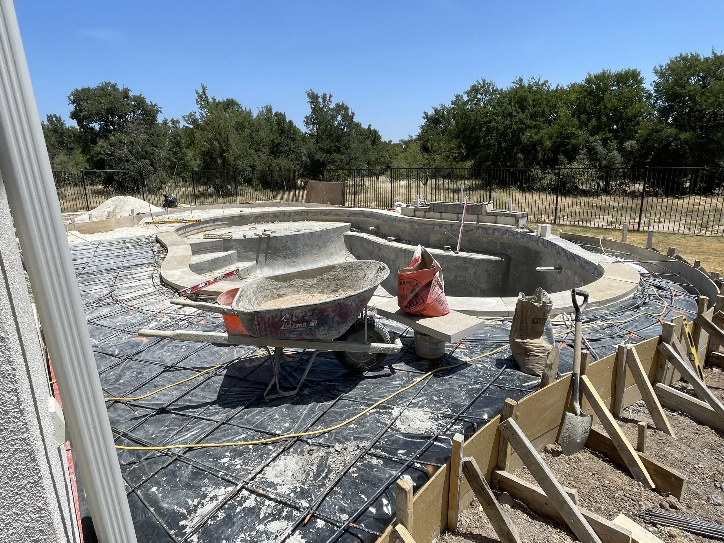 construction site of a swimming pool with concrete structure, red wheelbarrow, bags, and construction tools, surrounded by a wooden framework and safety fencing, with trees and blue sky in the background.