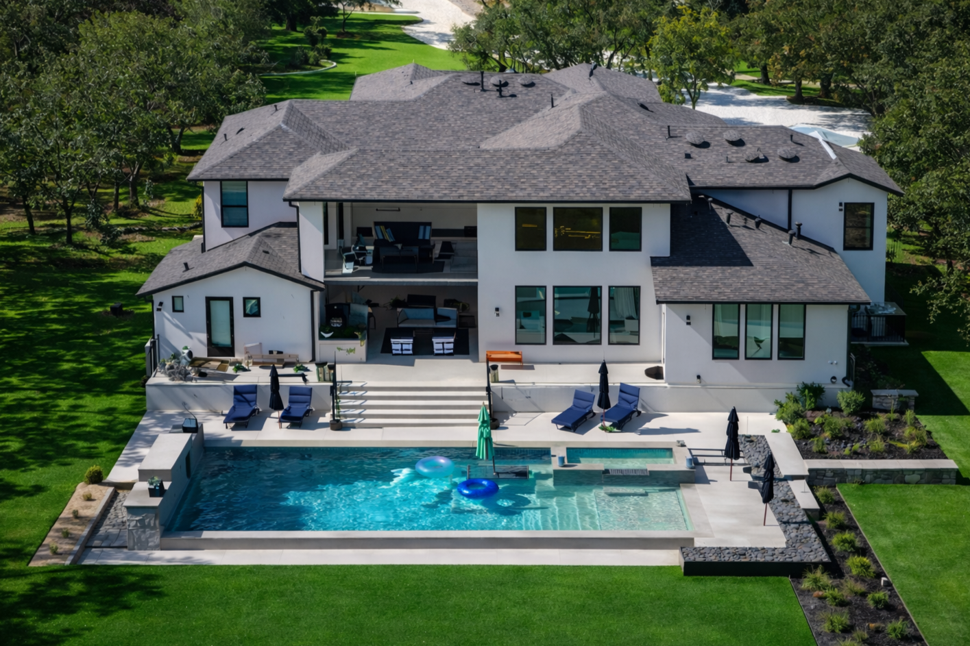 A large modern house with multiple levels and a dark gray roof, featuring large windows, with a backyard that includes a swimming pool, outdoor seating areas with umbrellas, and lush green lawn surrounded by trees.