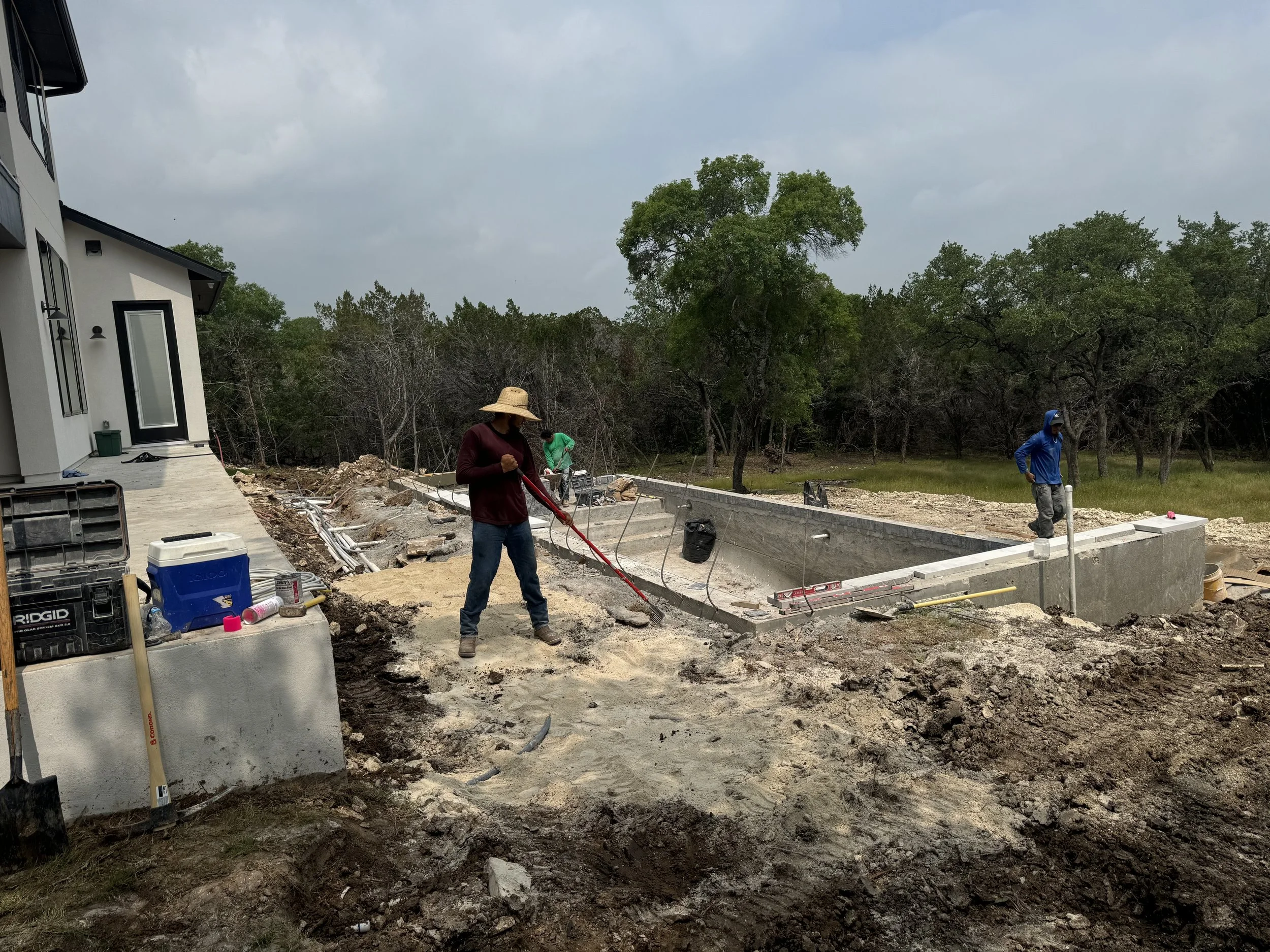 Construction workers building a swimming pool in a backyard, with trees in the background and construction tools around.