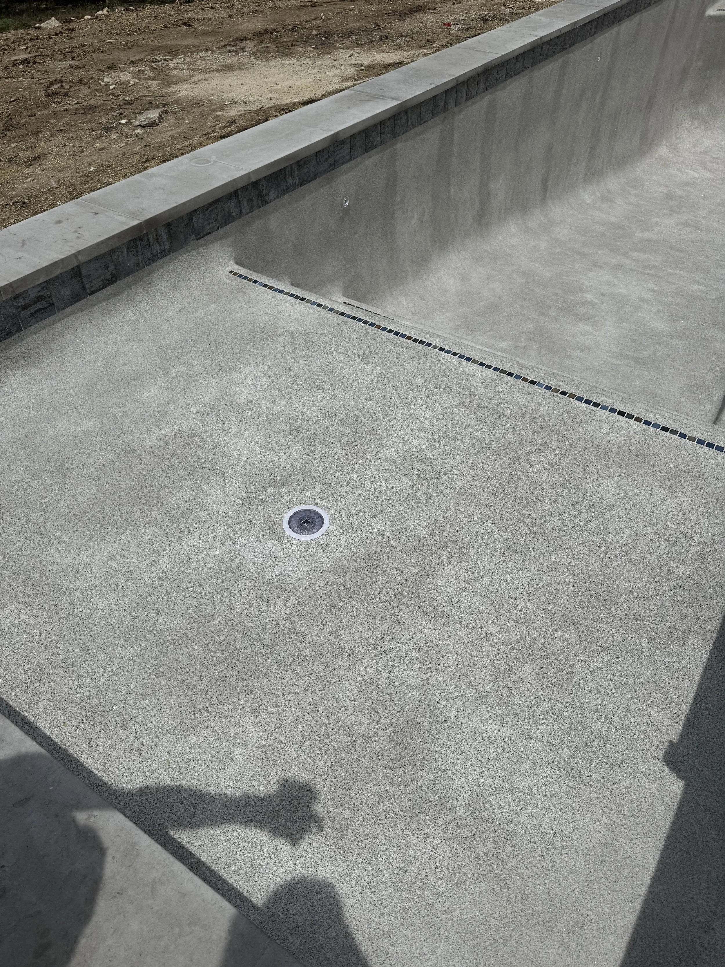 Empty outdoor concrete pool with drain, tile accents, and surrounding deck under construction.