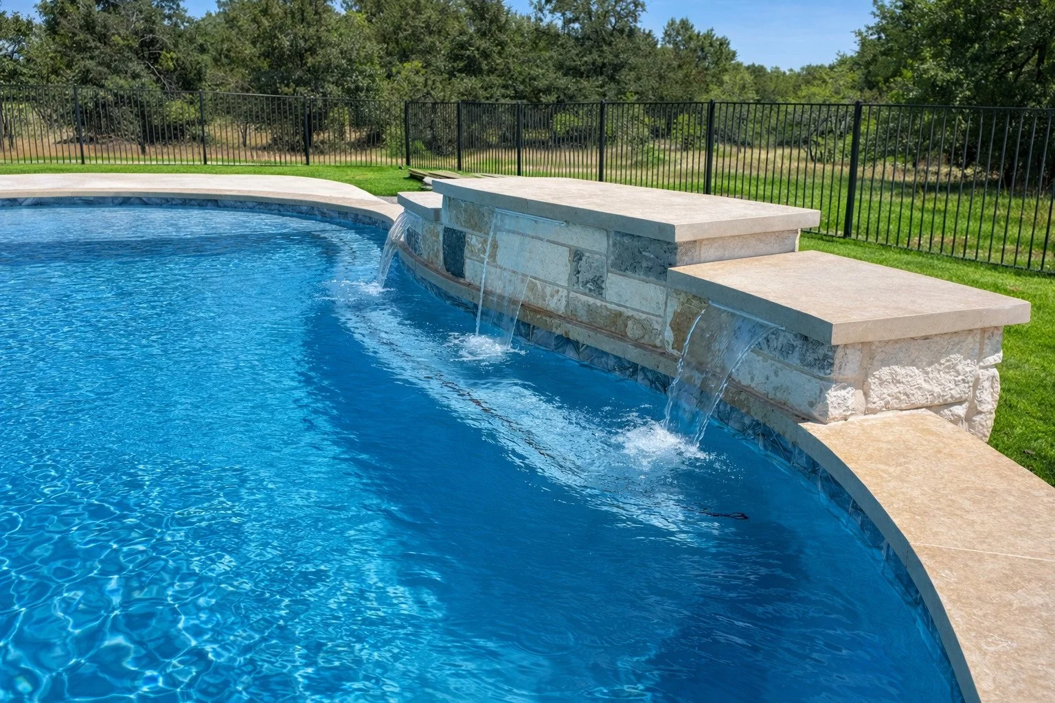 Swimming pool with a stone waterfall feature and a black metal fence surrounding it, with green grass and trees in the background.