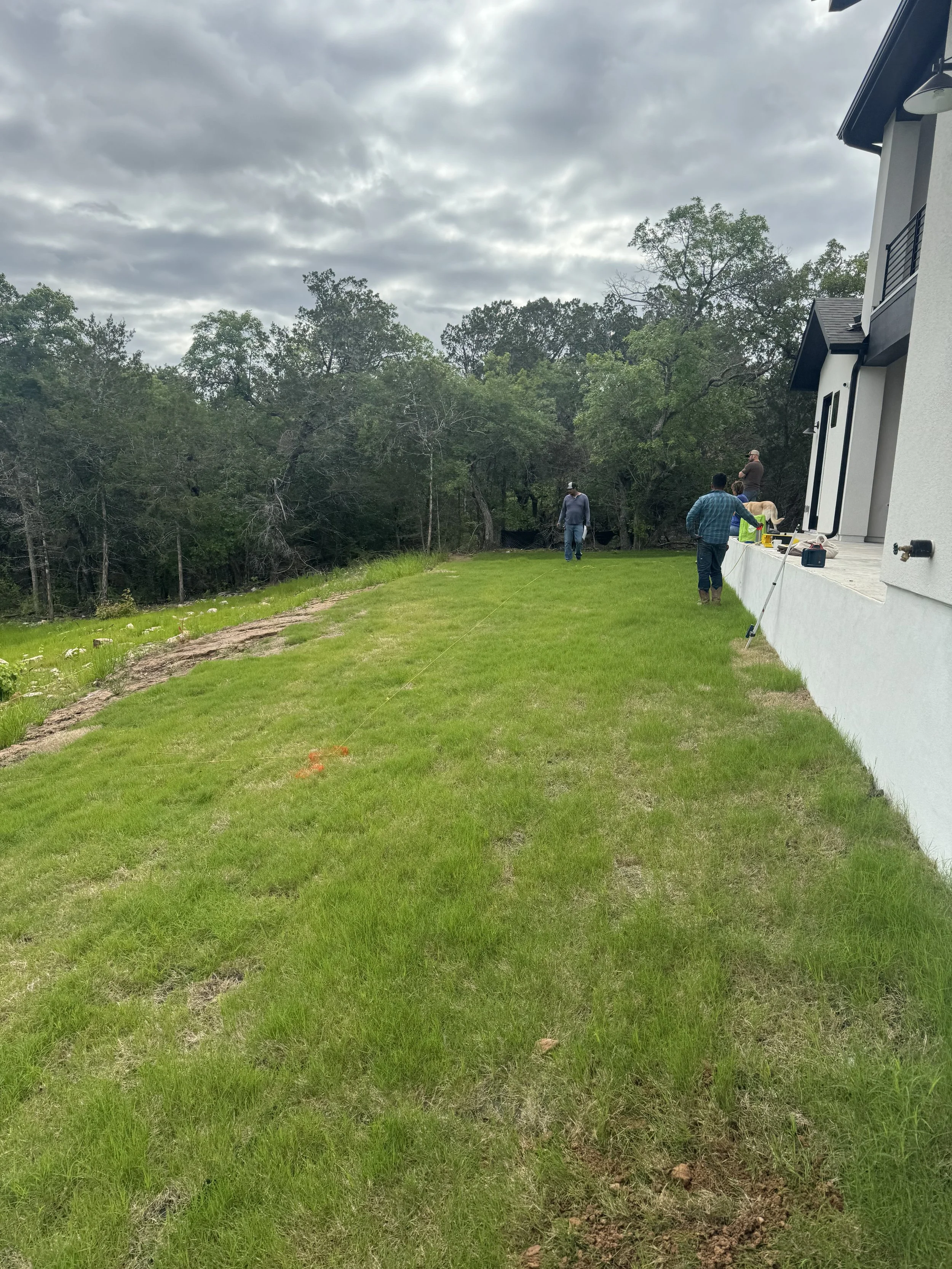 Group of people working on landscape installation or construction near the side of a modern house, with some tools and materials, green grassy yard, and trees in the background under a cloudy sky.
