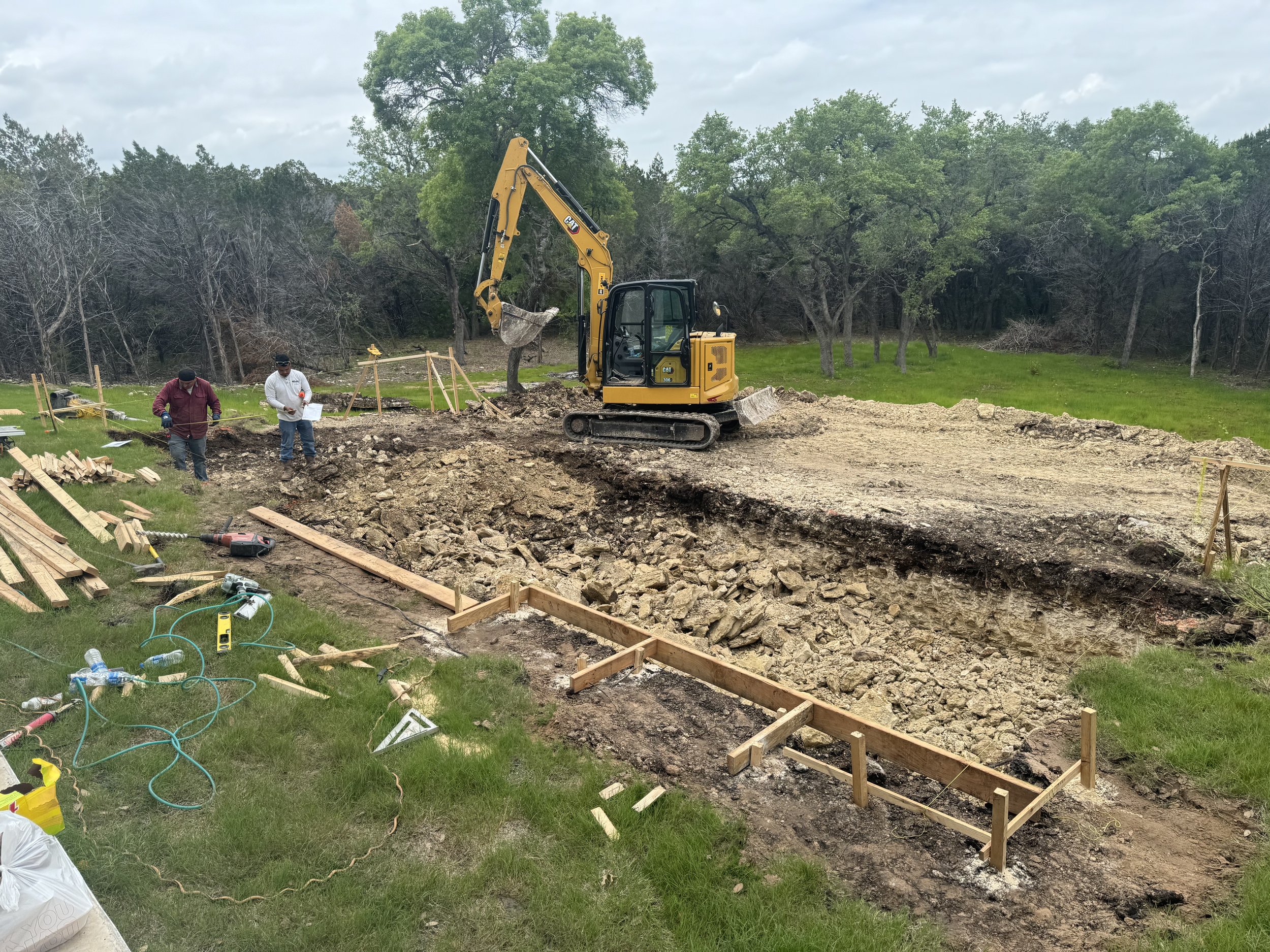 Construction site with a small excavator digging a large hole, two workers on site, raw earth and wooden framing, with tools and construction equipment scattered around.
