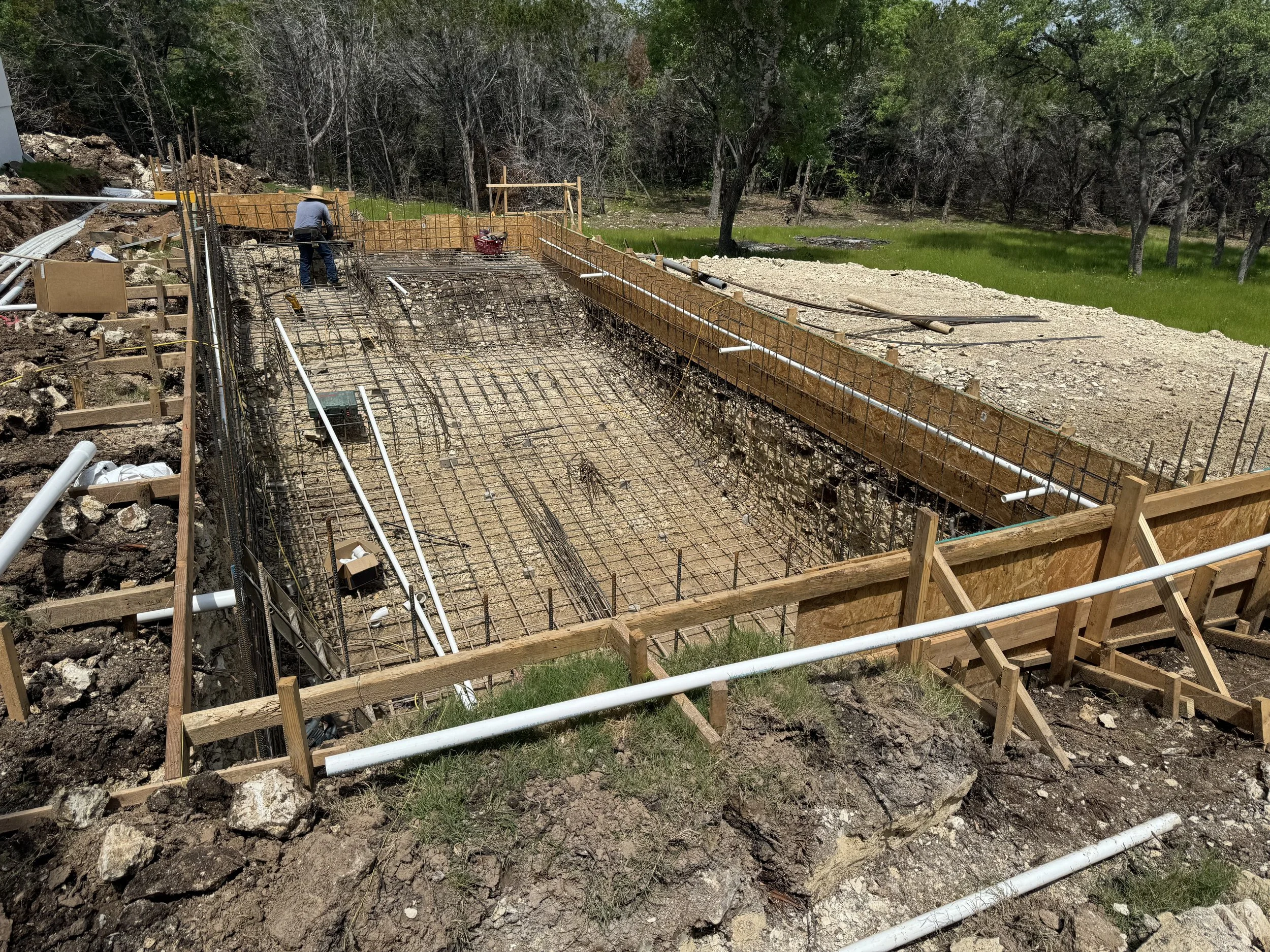 Construction site with rebar grid and wooden frames, worker in background, and pipes around the area, on a sloped hillside surrounded by trees and green grass.