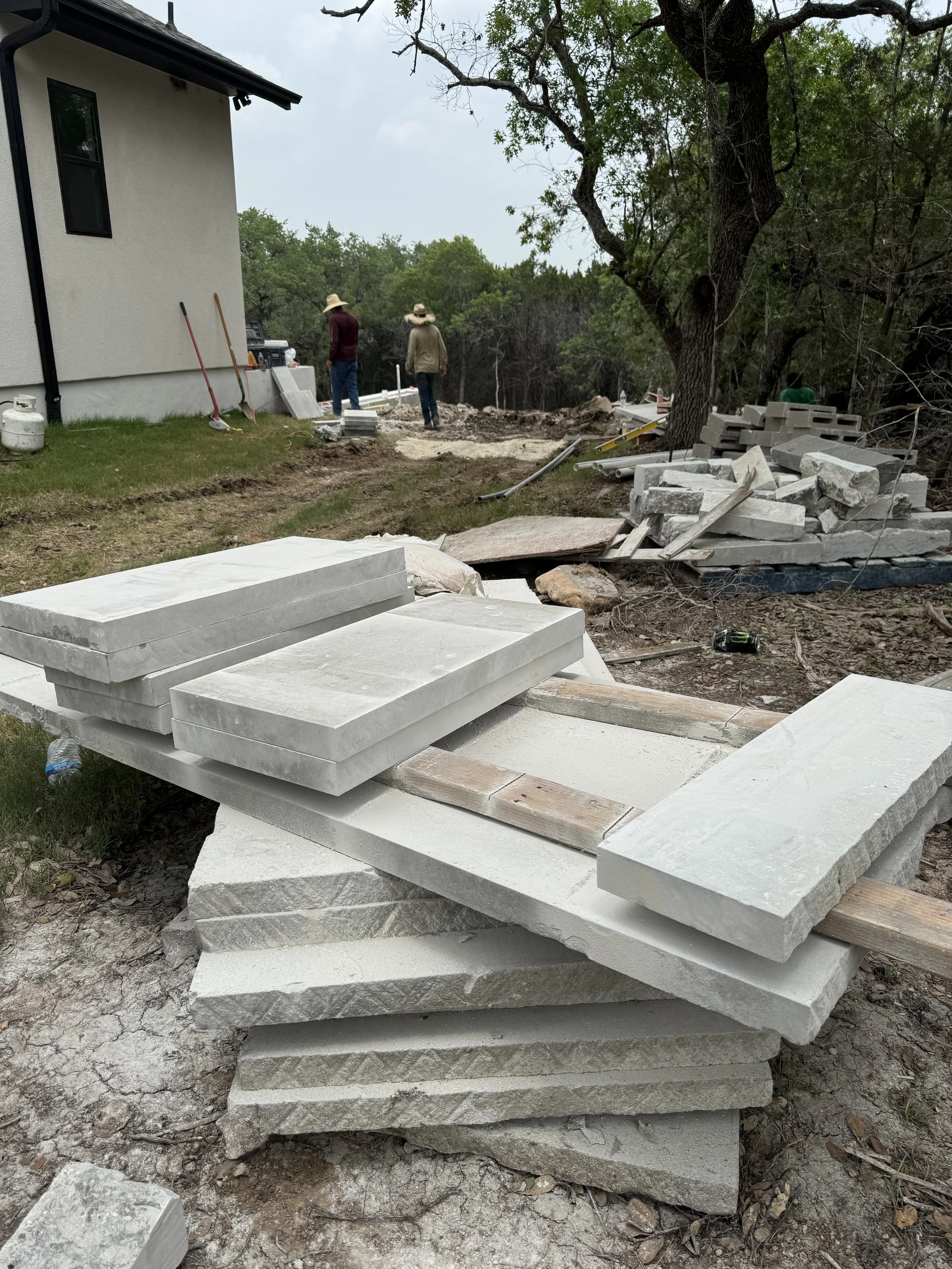 Construction site with stacked stone slabs and tools, two workers wearing straw hats working near a house, in a wooded area.