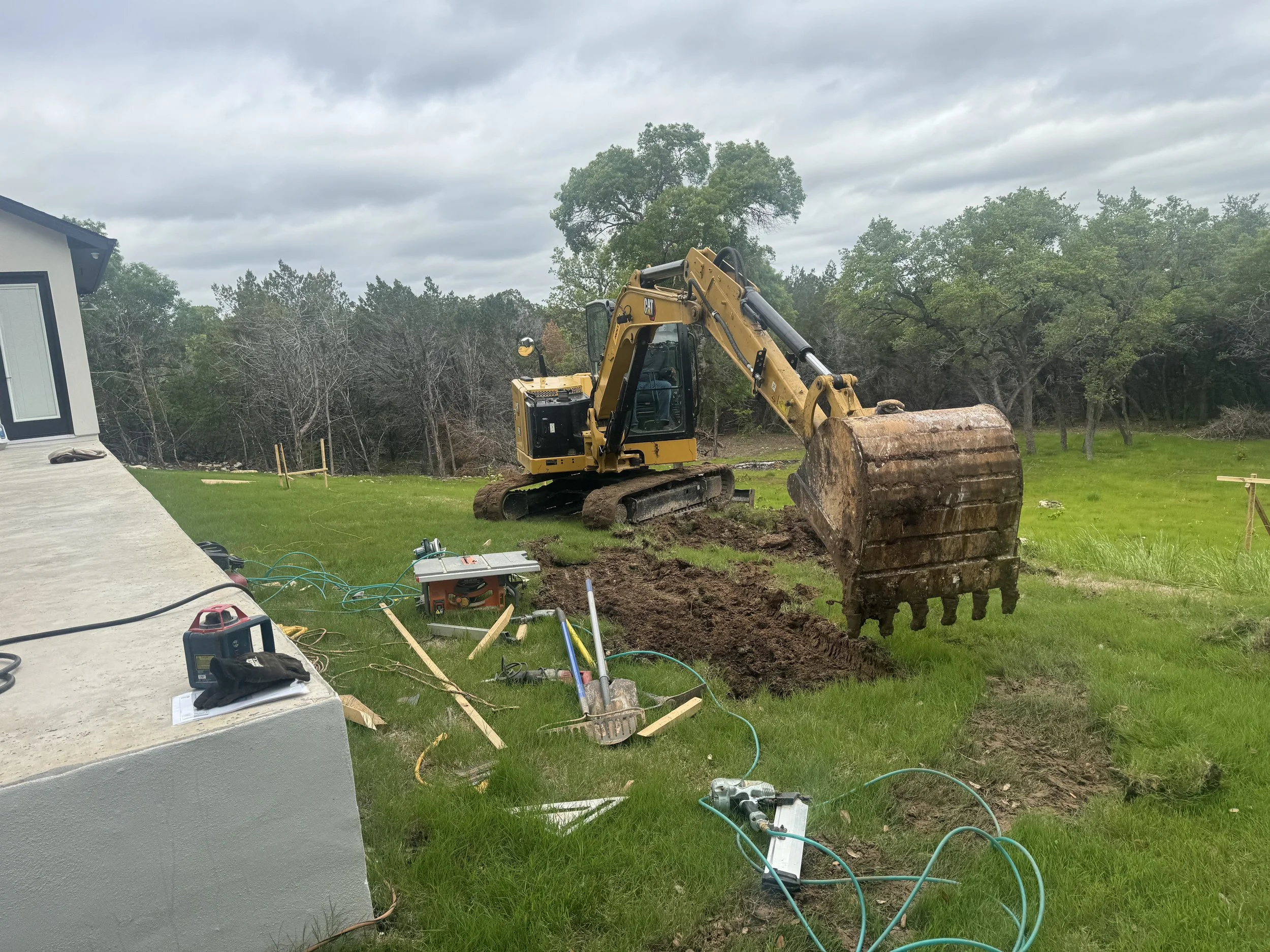 A yellow excavator digging a trench on a green lawn next to a house, construction tools and wires scattered on the ground, trees in the background under a cloudy sky.