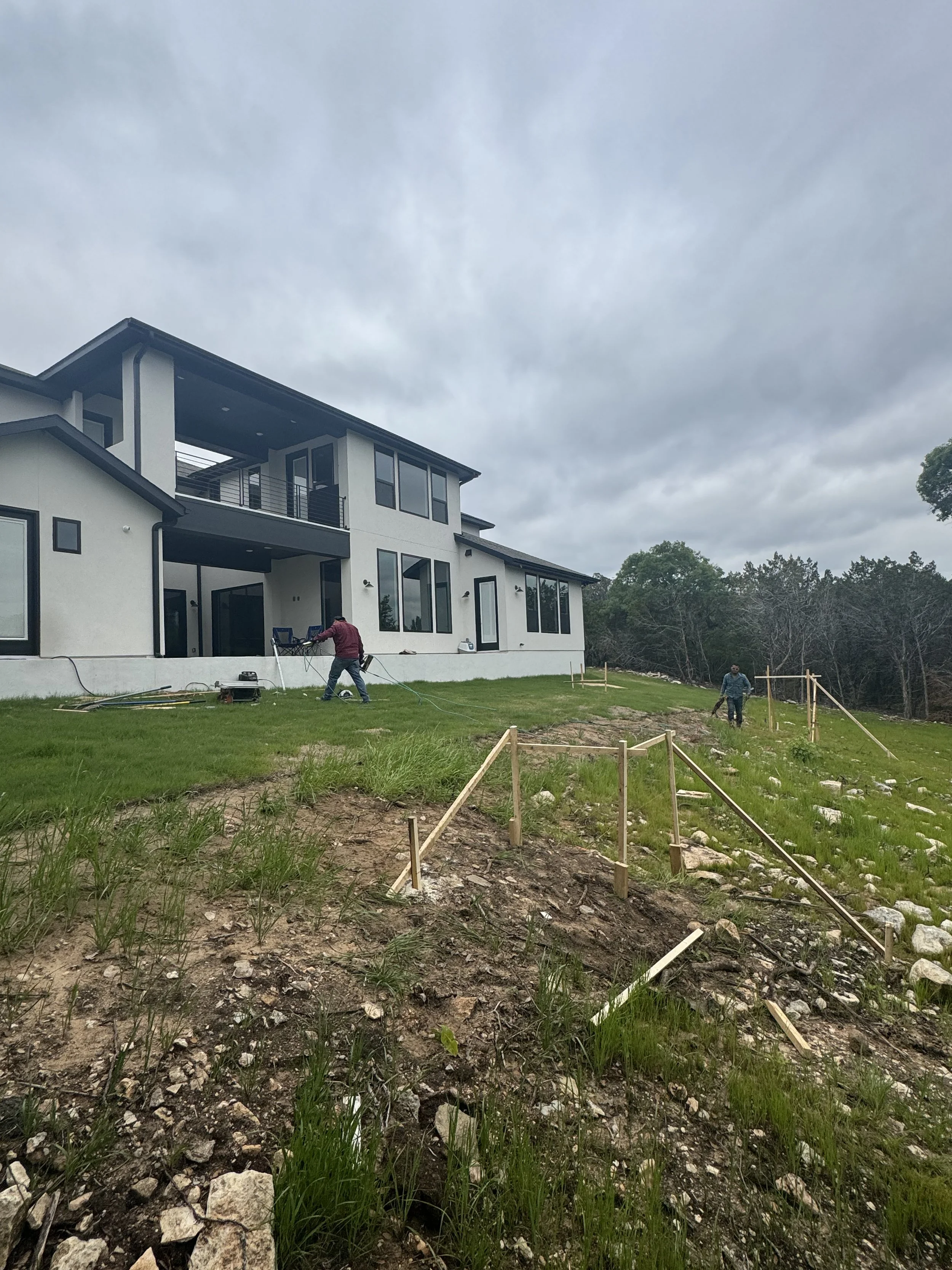 Construction workers installing or repairing a walking path outside a modern residential house with large windows and a balcony, on a cloudy day.