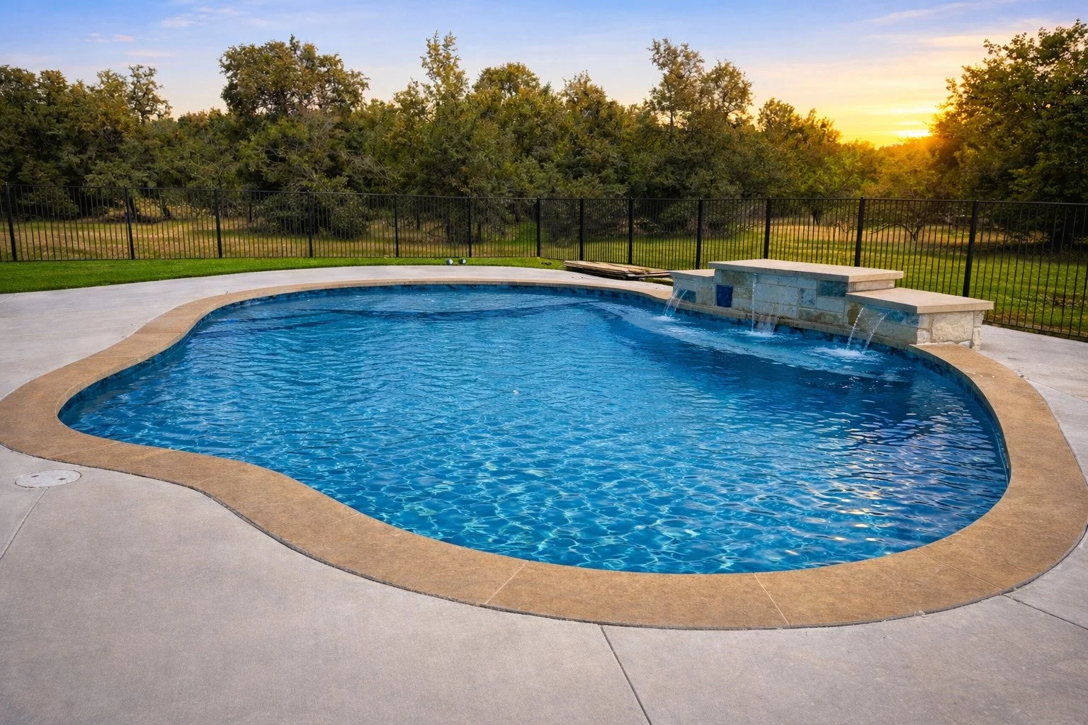 A backyard swimming pool with a waterfall feature, surrounded by a concrete deck and enclosed by a black metal fence. Trees and a sunset are visible in the background.