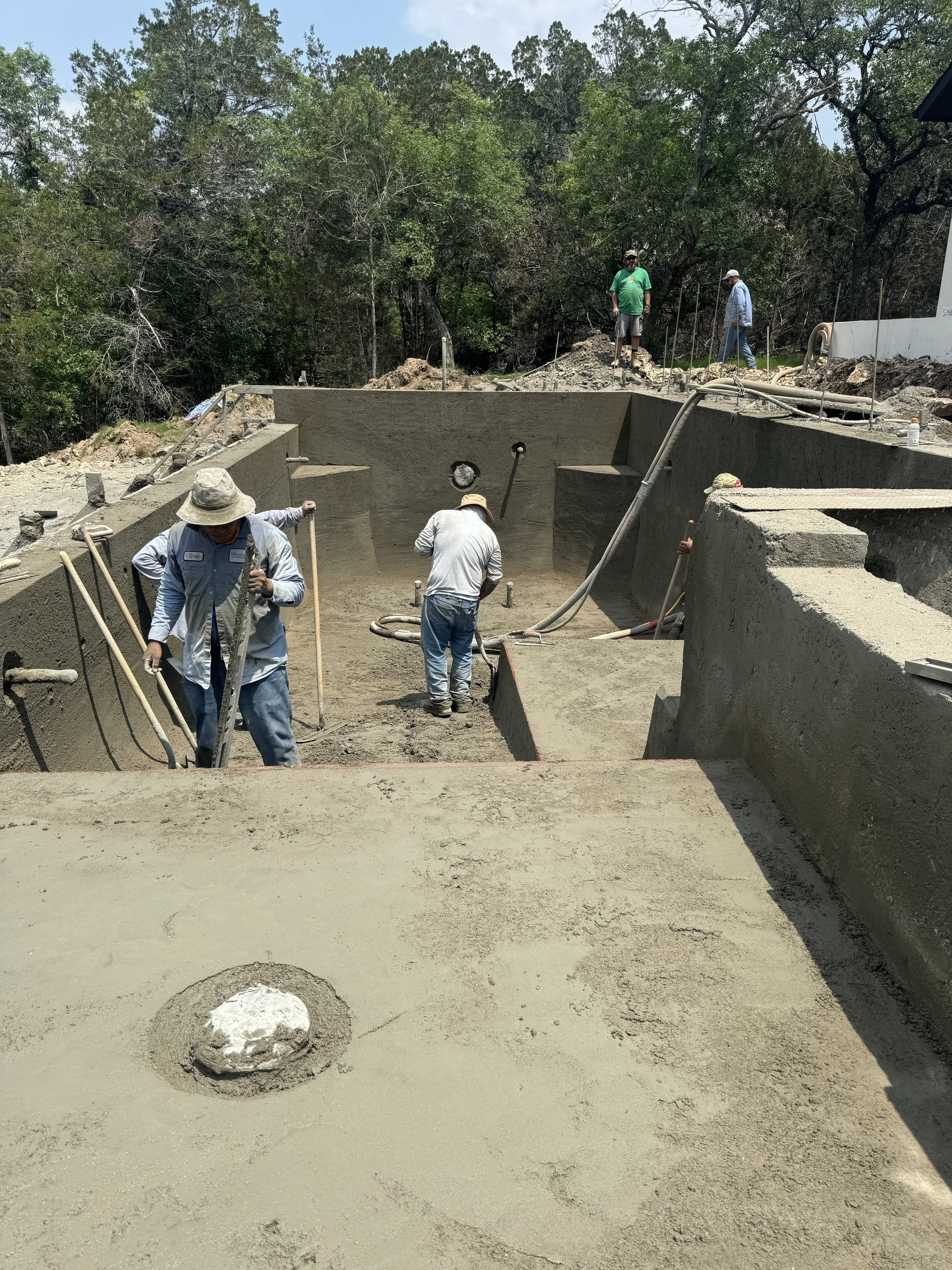 Construction workers are building a pool with concrete in a backyard, with some workers inside and others standing outside near a wooded area.