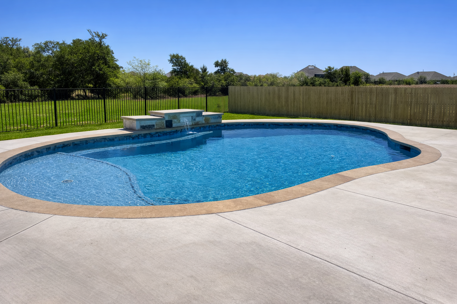 A clear blue in-ground swimming pool with a small round shallow section, surrounded by a concrete deck and a safety fence, with a grassy yard and houses in the background under a bright blue sky.