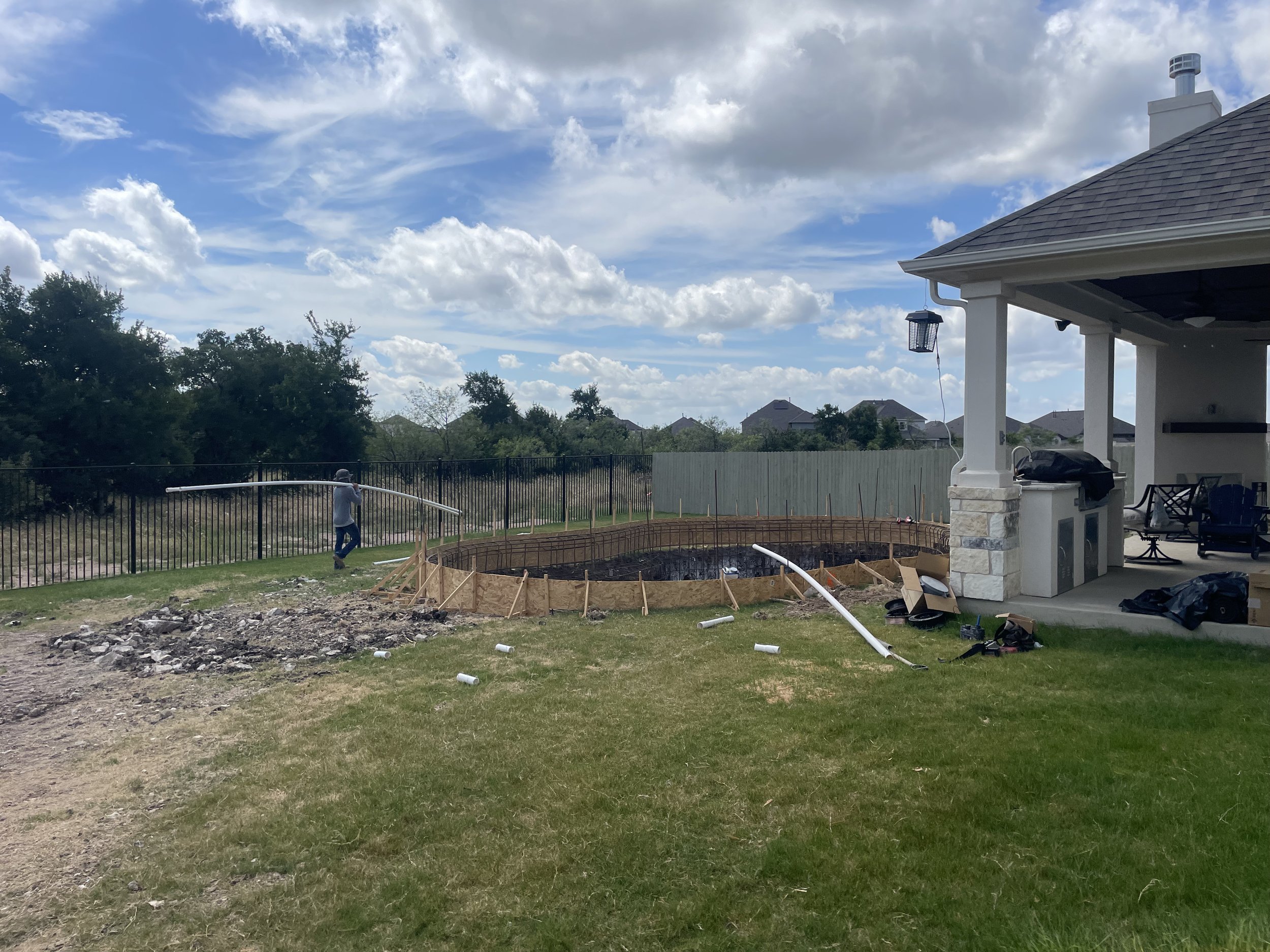 Construction site with a partially built in-ground pool, fencing, and a person working on the fence, on a bright day with cloudy sky.