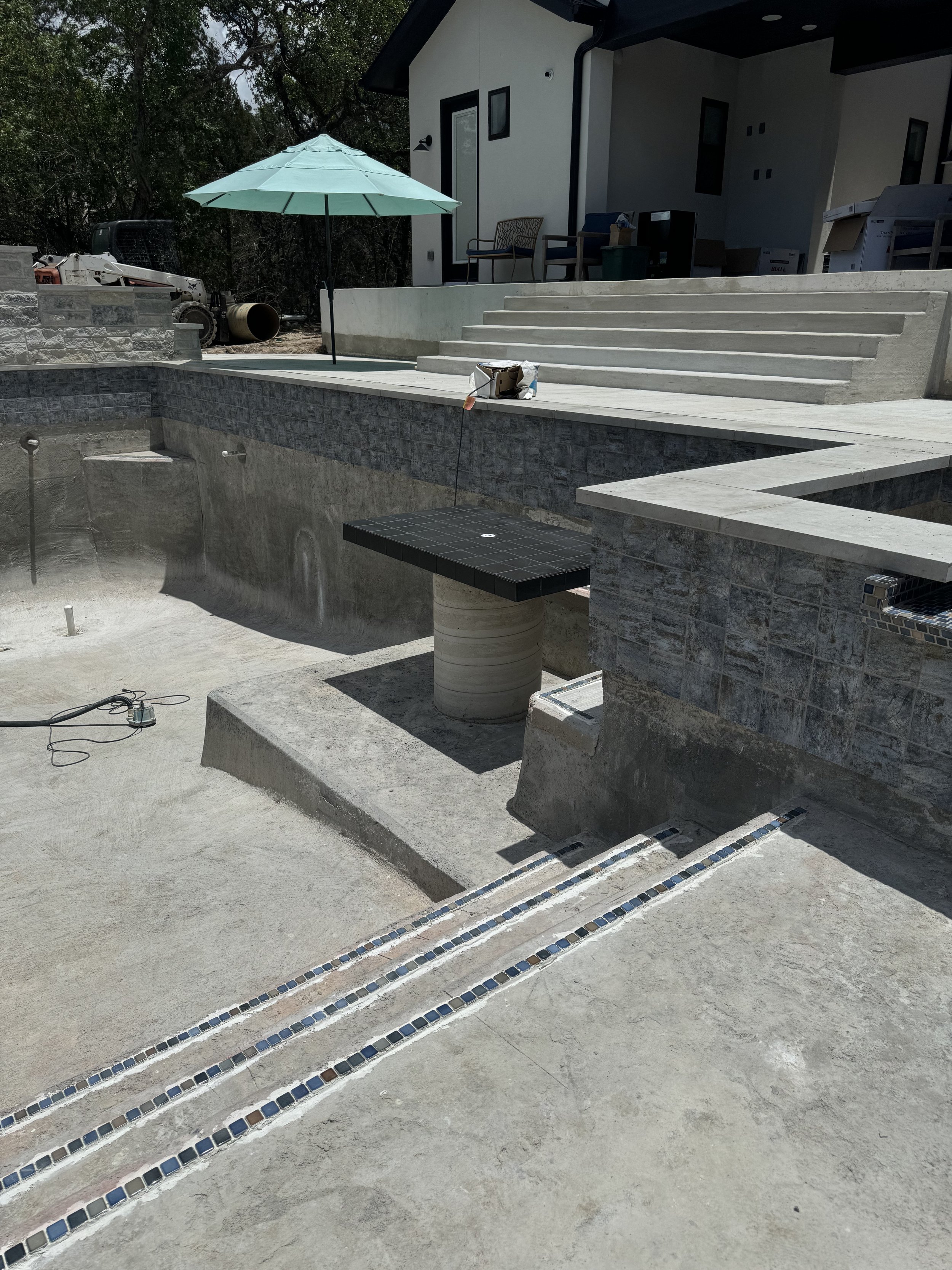 Under construction or renovation backyard with a pool area, partially built, with a small table with black tiles, a green umbrella, some construction tools, and a house with stairs leading up to a porch.