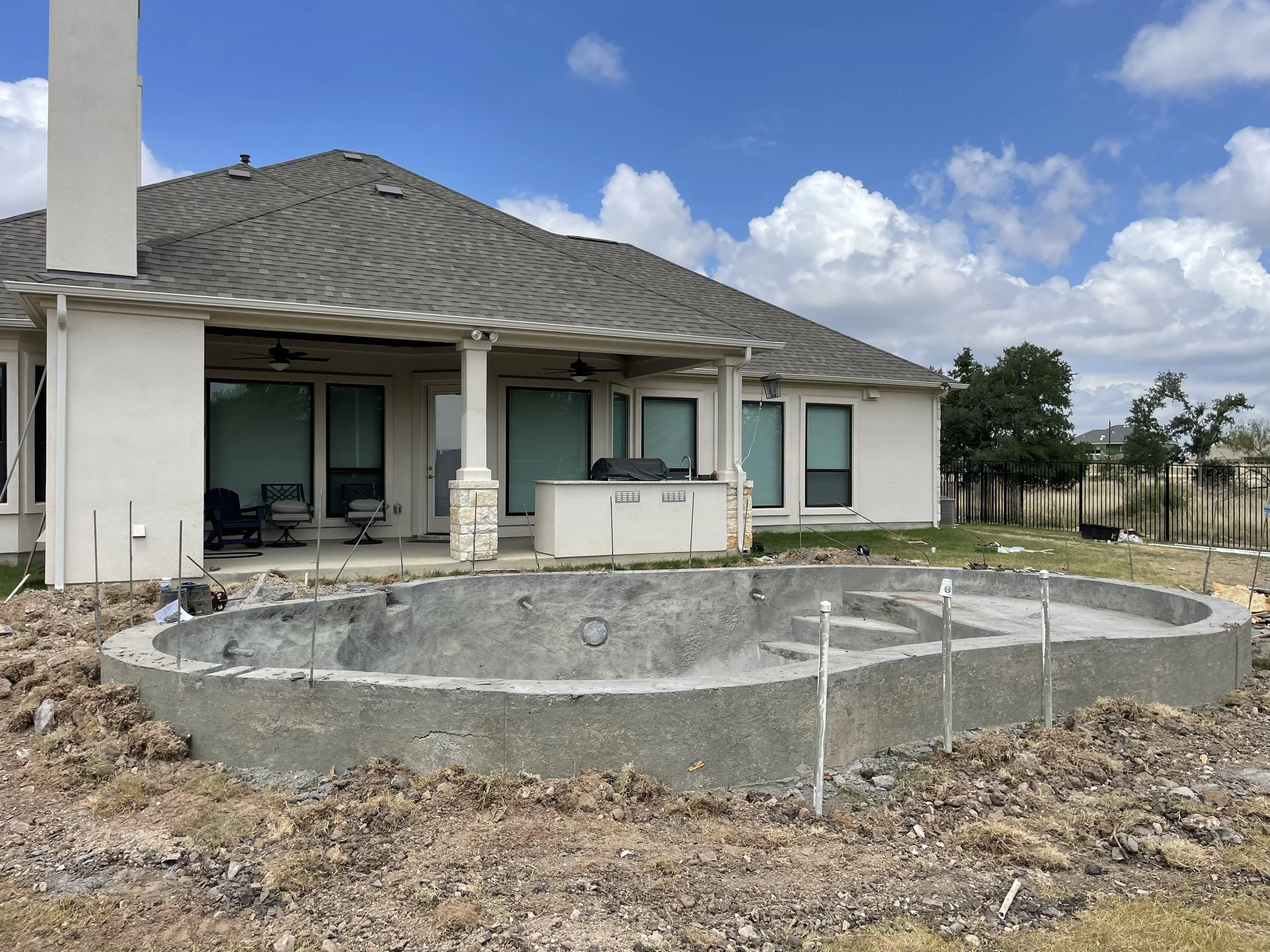 Backyard of a house with an empty, unfinished swimming pool in the foreground, surrounded by construction dirt and debris, and a covered patio with seating and ceiling fans in the background.