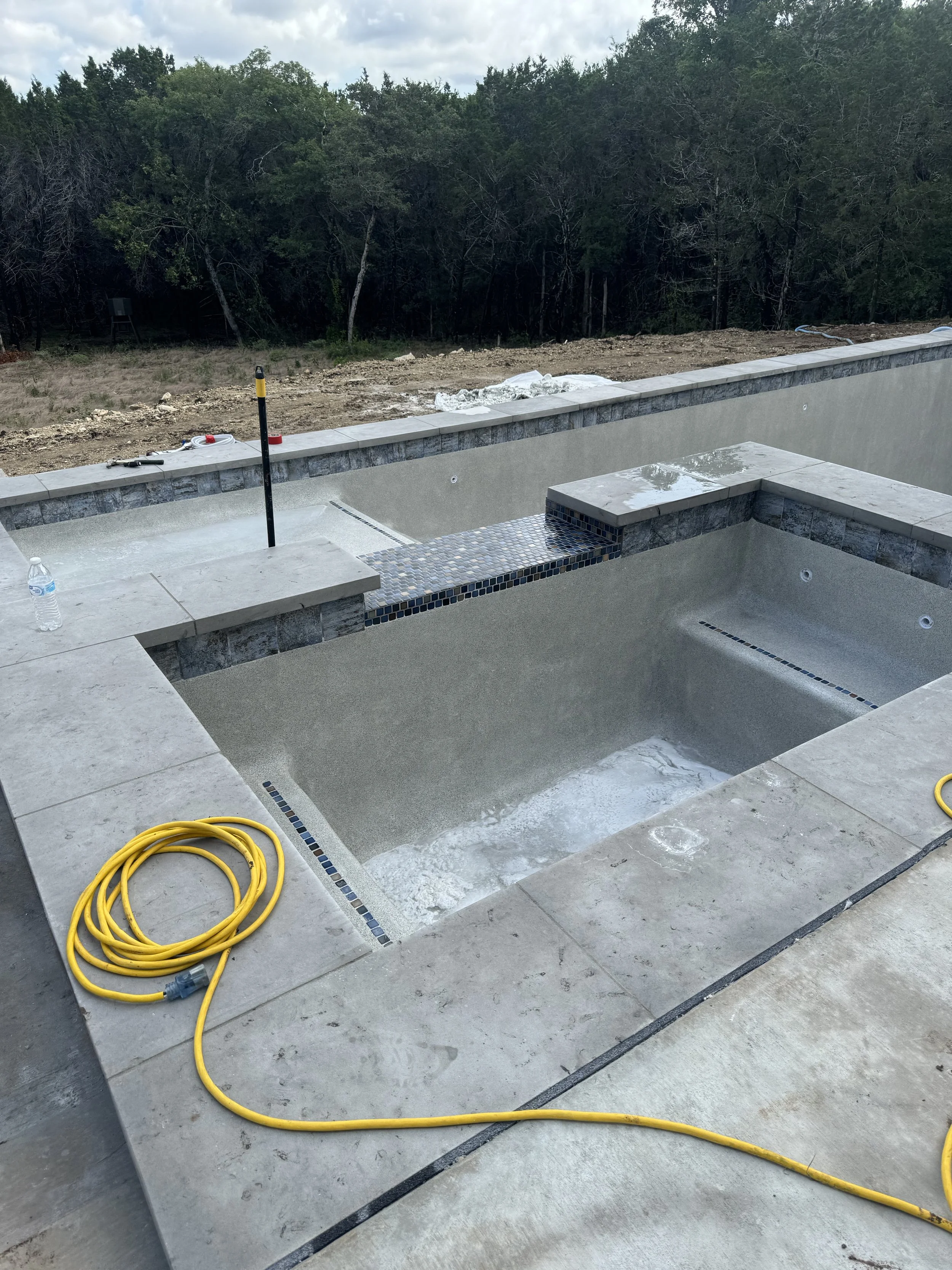 An empty backyard swimming pool under construction, with construction tools and supplies around it, and a wooded area in the background.