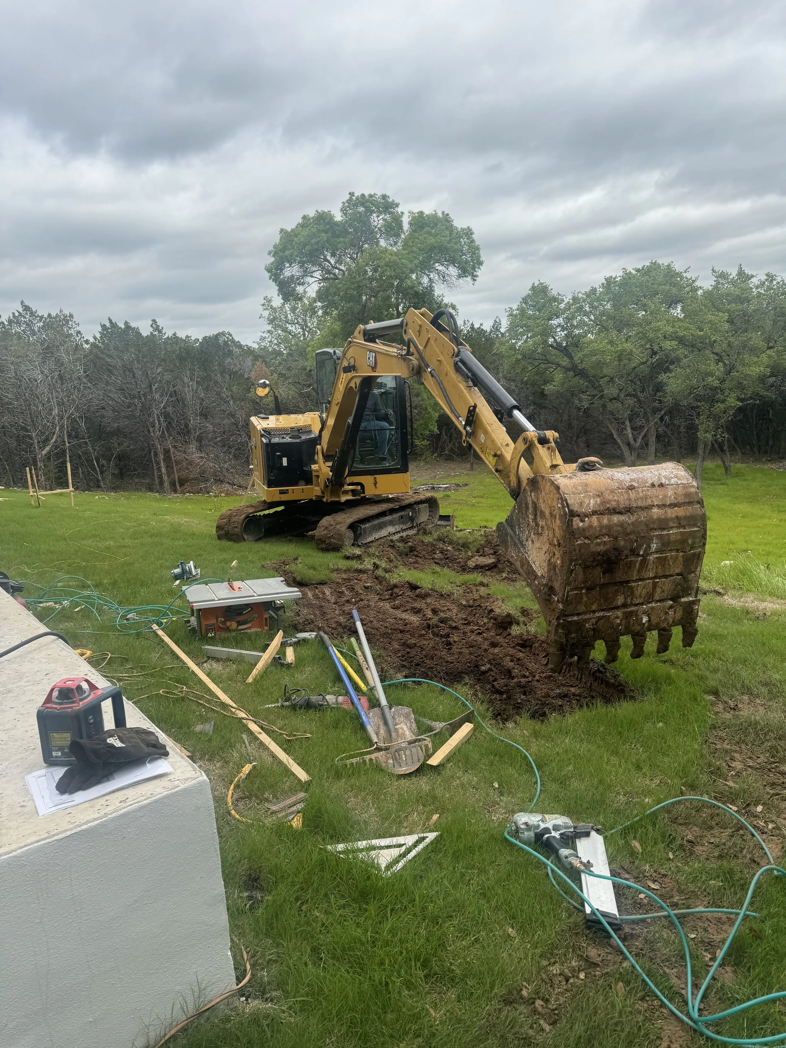 A yellow bulldozer excavating soil on a grassy field with trees in the background, construction tools and equipment in the foreground.