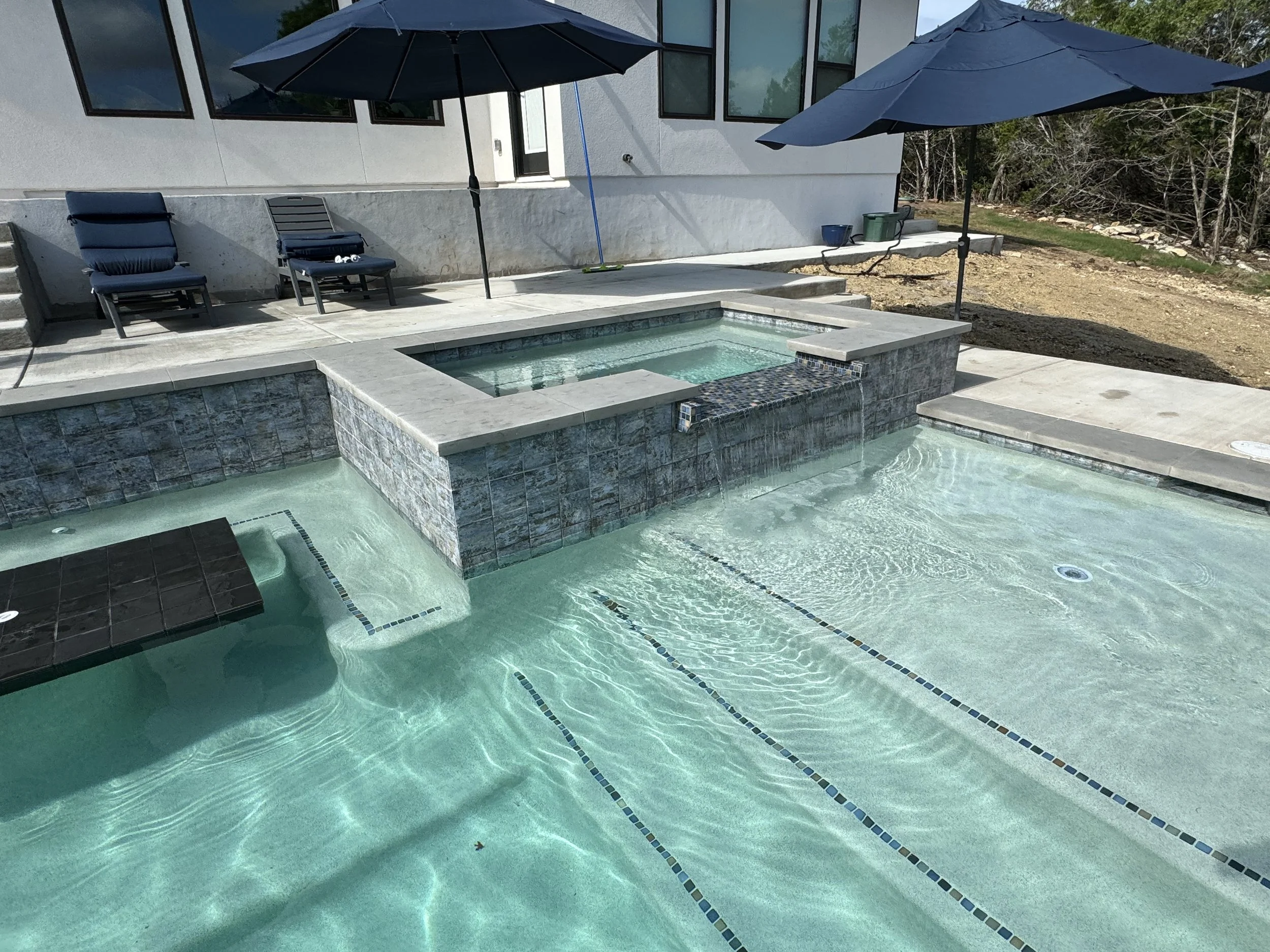 House with an outdoor pool and hot tub, shaded by large umbrellas, with lounge chairs and a patio area.