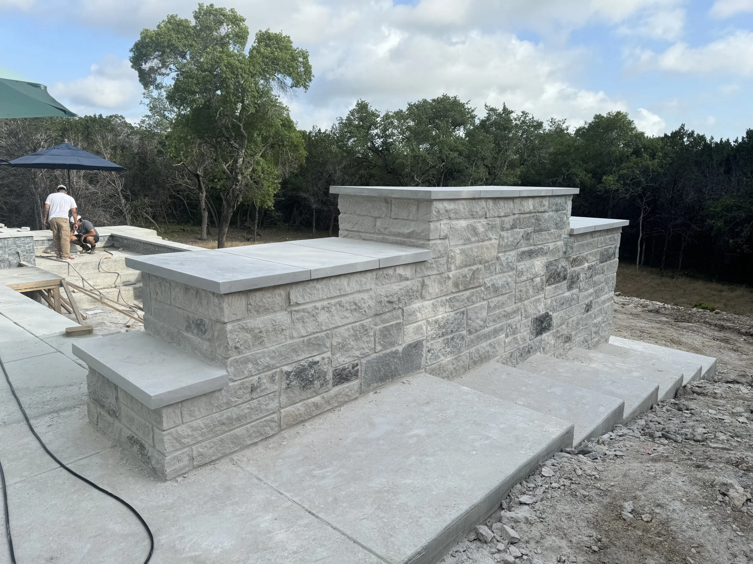 Construction workers building a stone and concrete structure outdoors on a construction site with trees and a partly cloudy sky in the background.