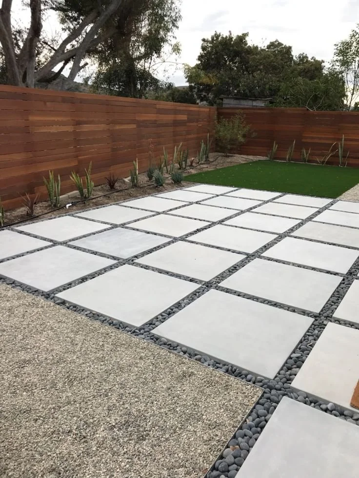 A backyard with a wooden privacy fence, landscaped with small plants along the fence, a patch of artificial grass, and a patio area with large square concrete pavers separated by gravel.