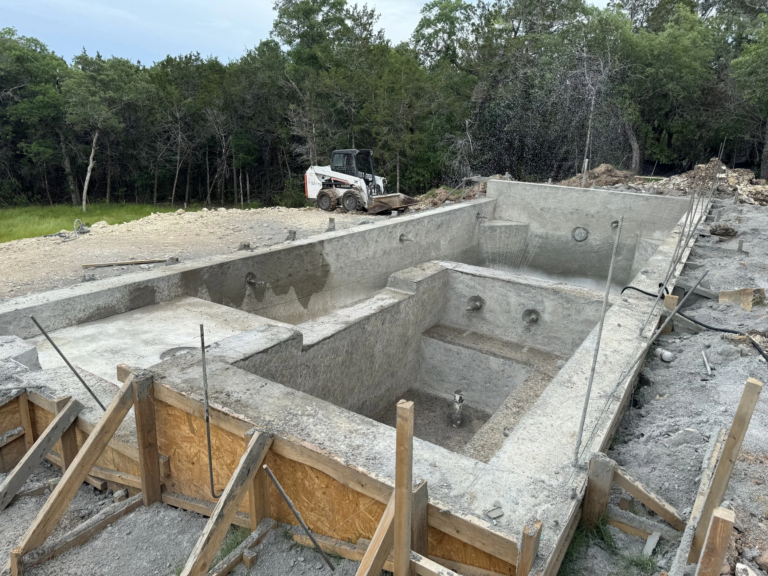Under construction hot tub with wooden framing, set on rocky ground with construction equipment in the background, surrounded by trees.
