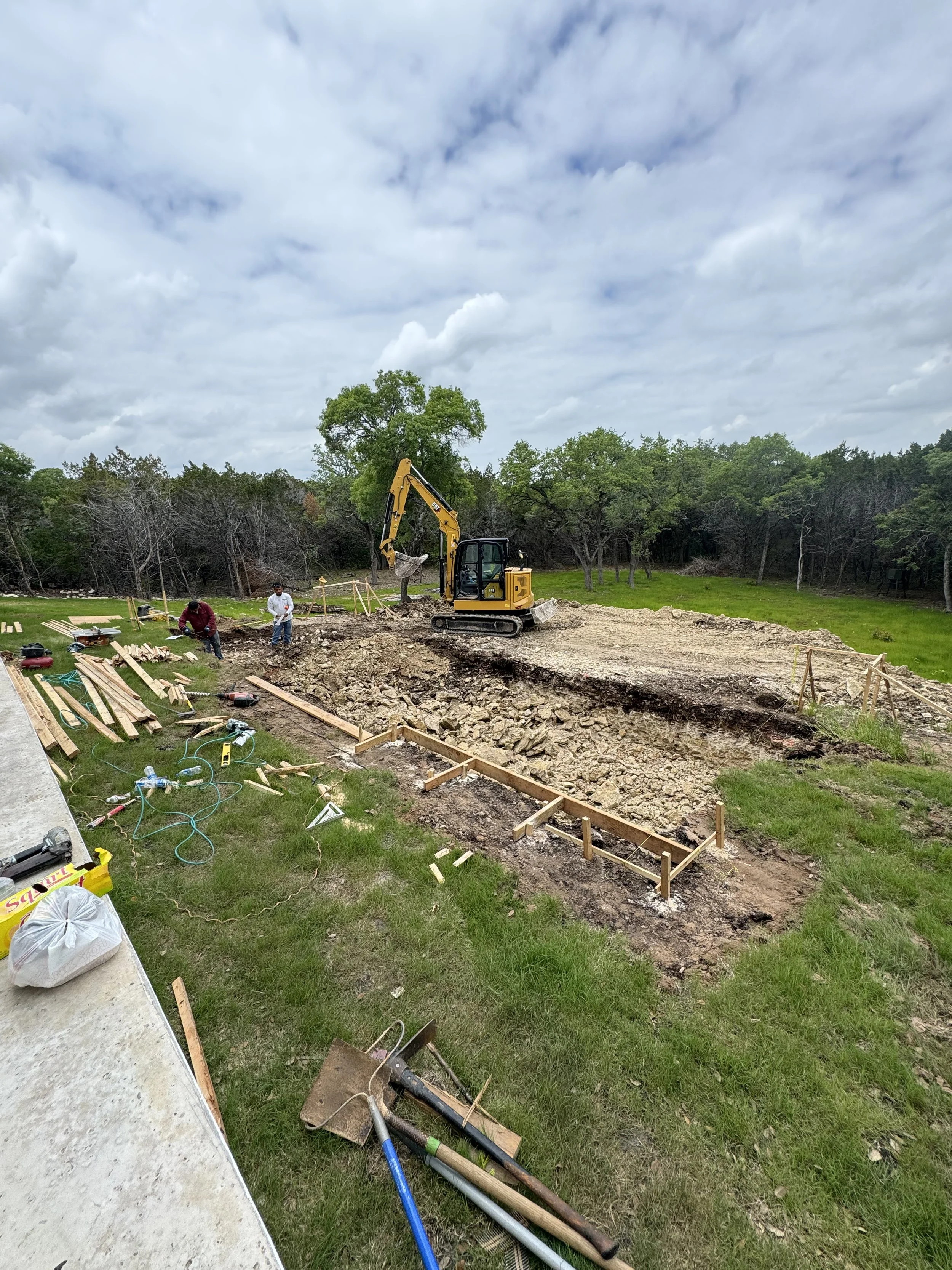 Construction site with a yellow excavator and workers digging a trench in a grassy area surrounded by trees under a cloudy sky.