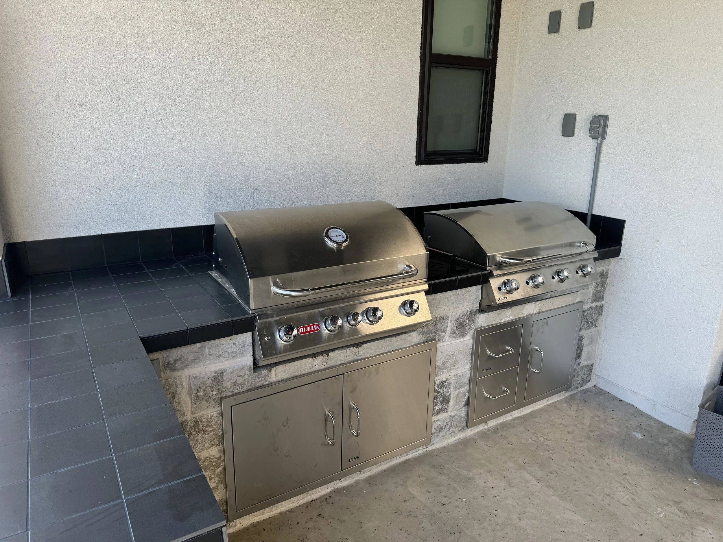 Two stainless steel outdoor grills with temperature gauges. One grill is branded 'Bull.' The grills are installed on a brick counter with black tiled countertop. Behind the grills, there is a small window and some electrical outlets on the white wall