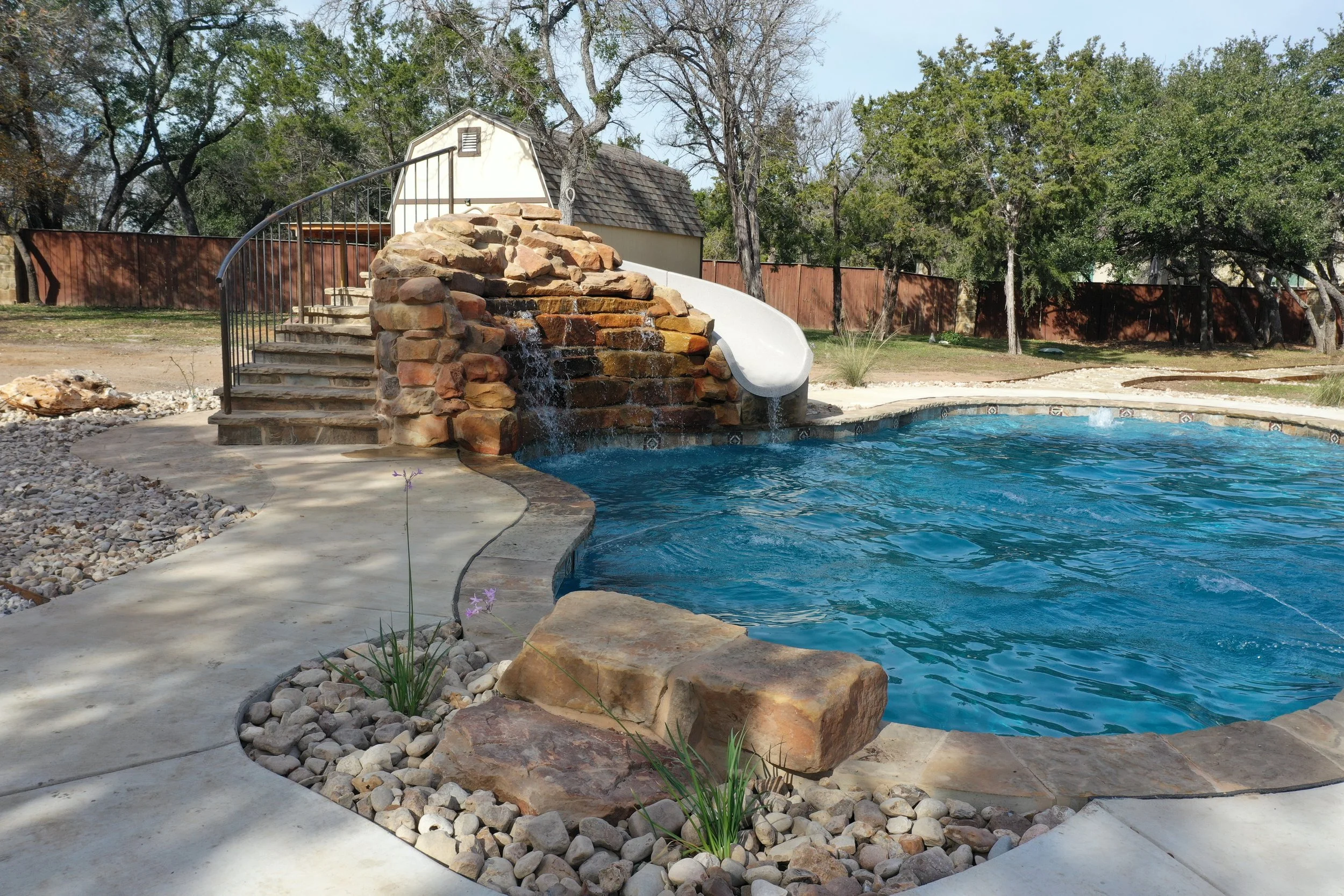 Residential backyard with a swimming pool featuring a stone waterfall and water slide, surrounded by a concrete patio and a wooden fence, with trees in the background.