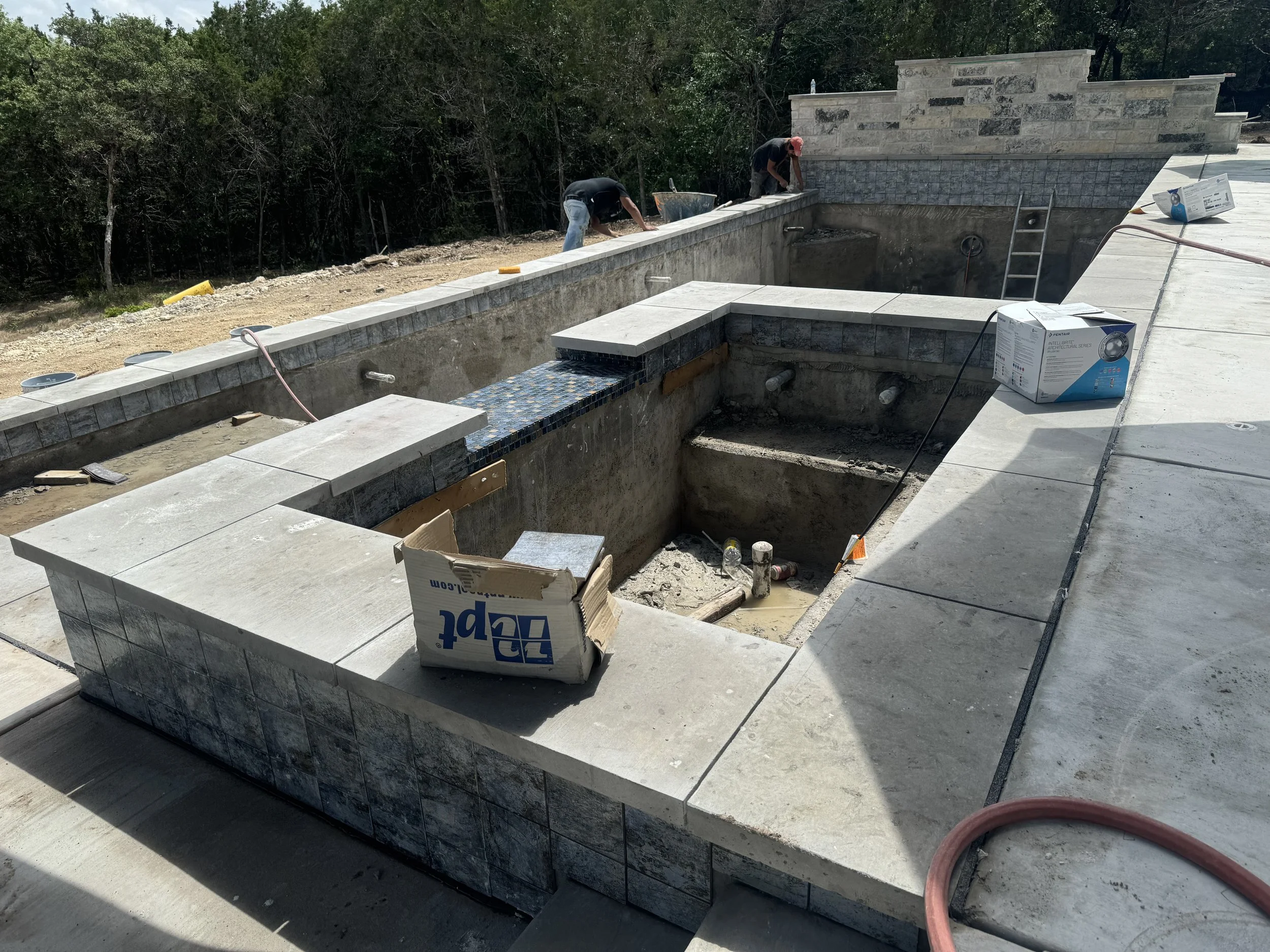 Construction workers working on building a pool with tiled edge, with walls and steps partially finished, and construction materials scattered around.