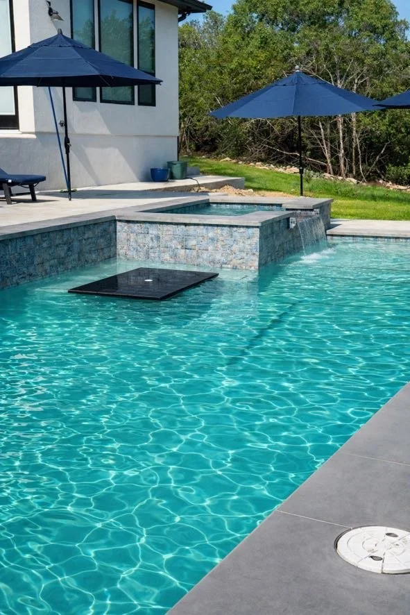 A backyard swimming pool with a hot tub, blue umbrellas, and a modern house with large windows.
