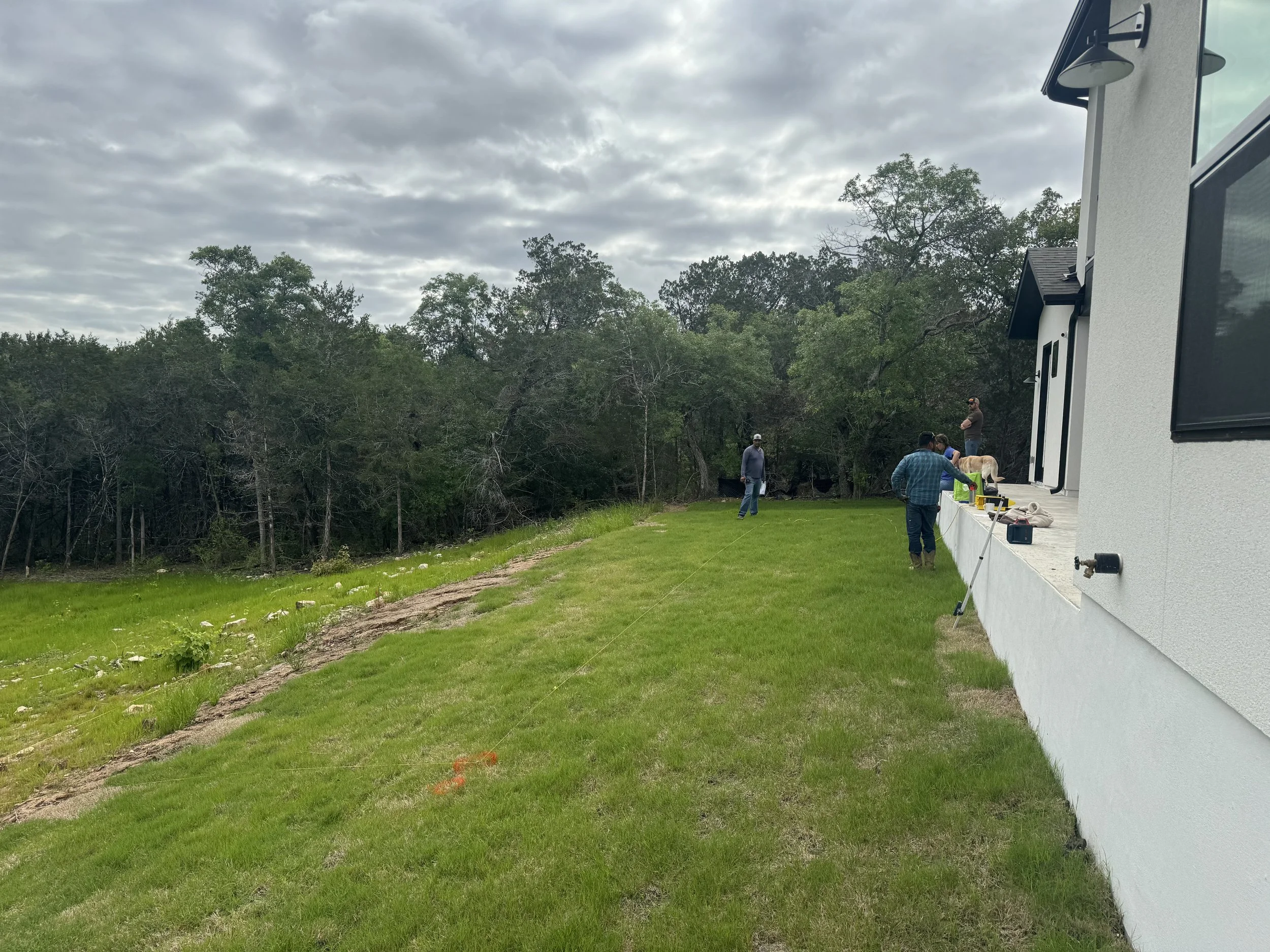 A group of people working outdoors on a lawn beside a white residential building, with a forested area in the background and cloudy sky overhead.