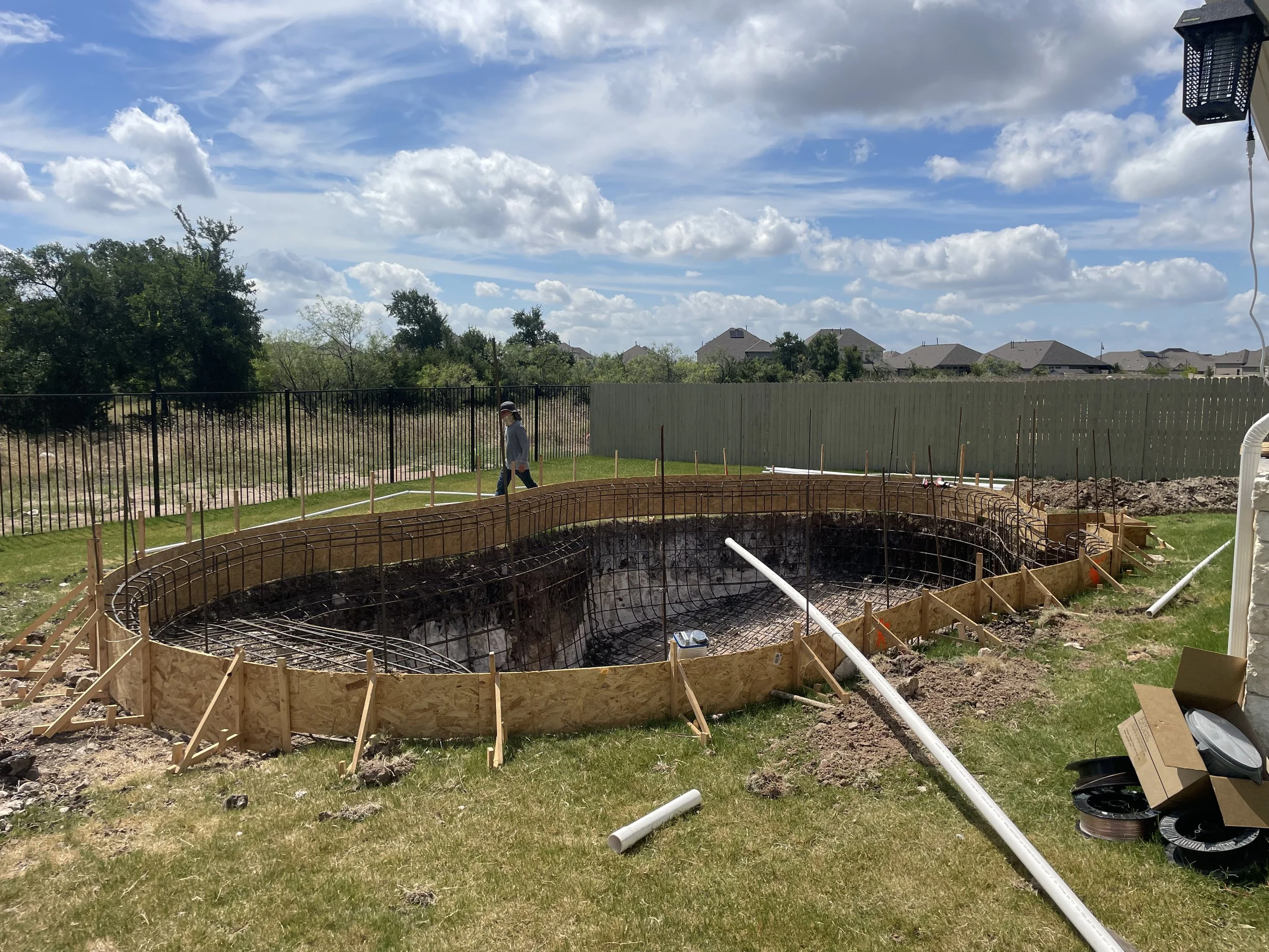 Construction site of a small pool with wooden and metal framing, surrounded by green grass and a backyard fence, under a partly cloudy sky.