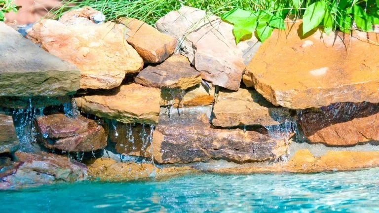 Rock formation with small waterfalls beside a body of water and green foliage at the top edge.