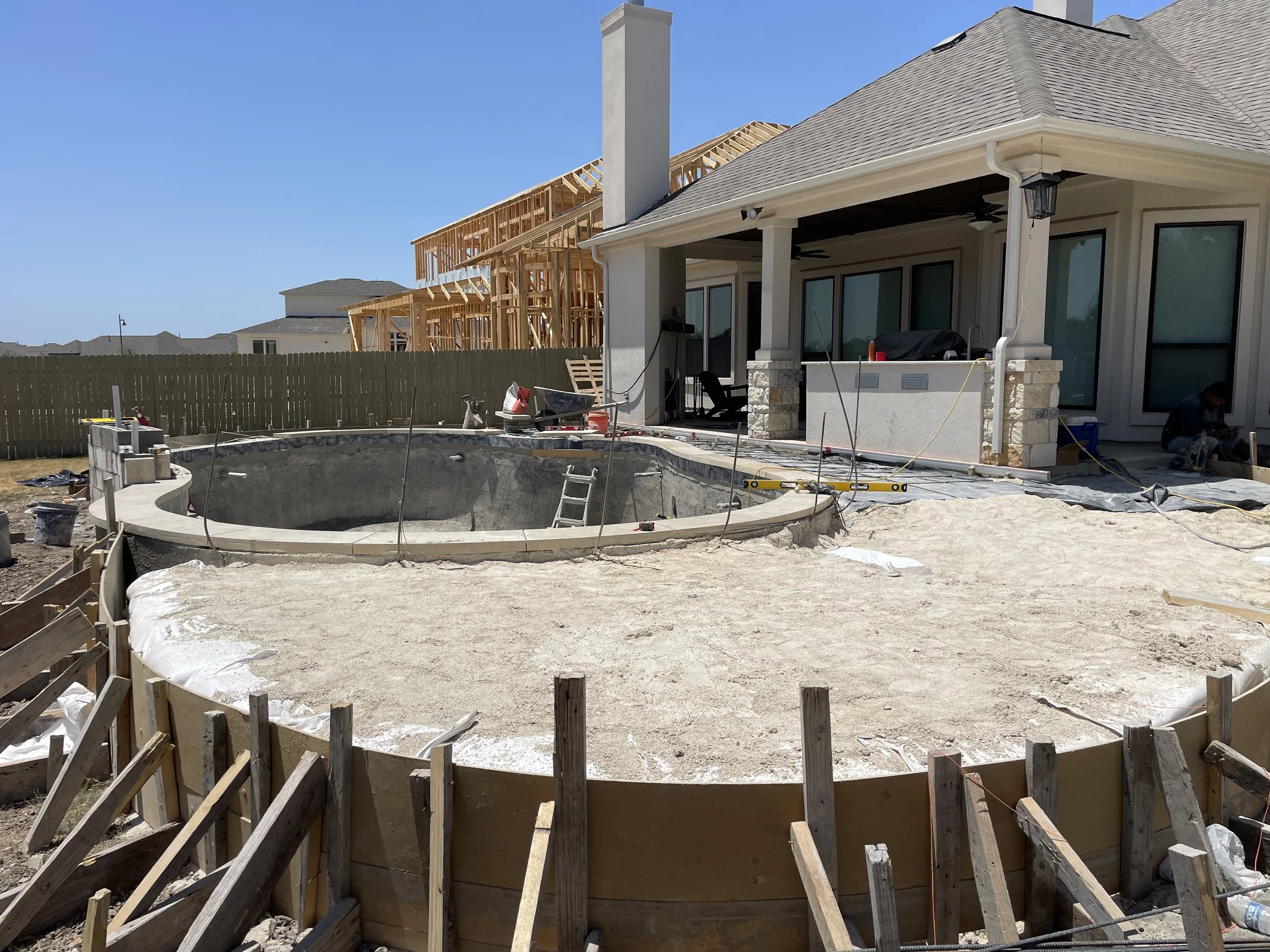 Construction site of a backyard swimming pool with a concrete shell partially built, surrounded by wooden forms, with a ladder inside. There is a house in the background under renovation, with visible wooden framing. Two workers are present on the ri