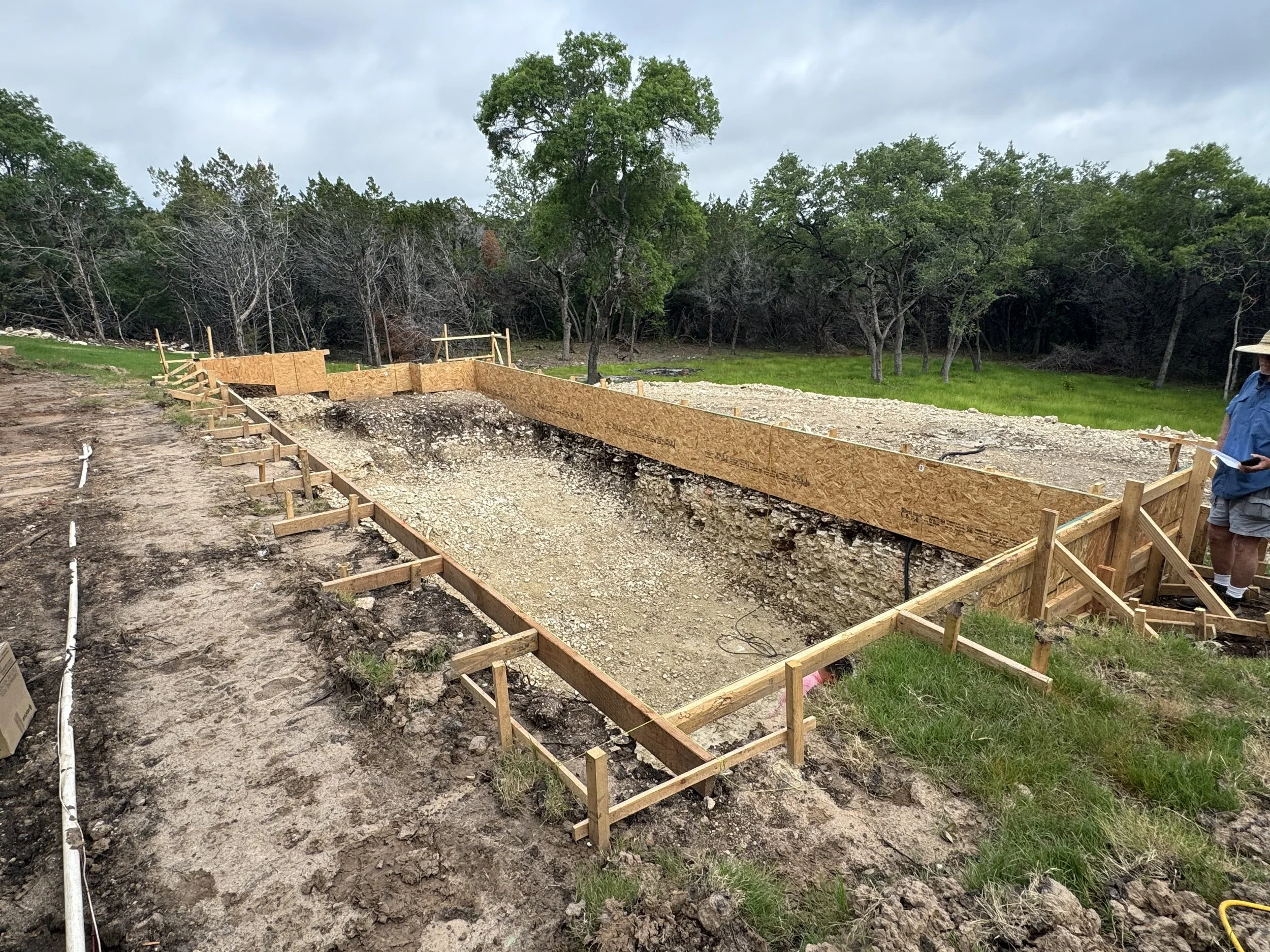 Construction site with a wooden frame for a foundation in progress, surrounded by dirt, gravel, and trees in the background, with a person holding a tablet on the right.