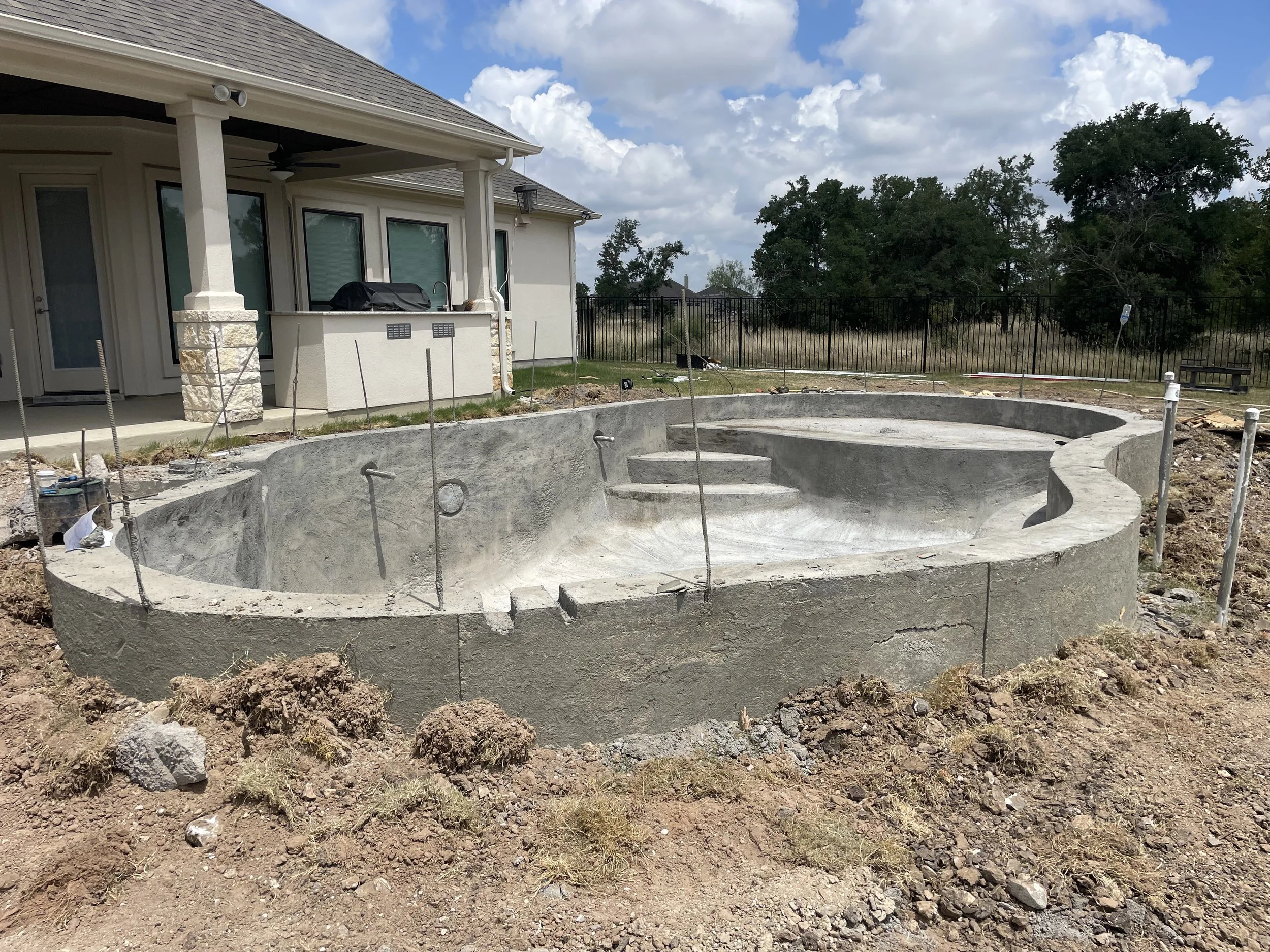 Unfinished in-ground swimming pool under construction in backyard with dirt surrounding it, next to a house with a covered patio, trees, a cloudy sky, and fencing in the background.