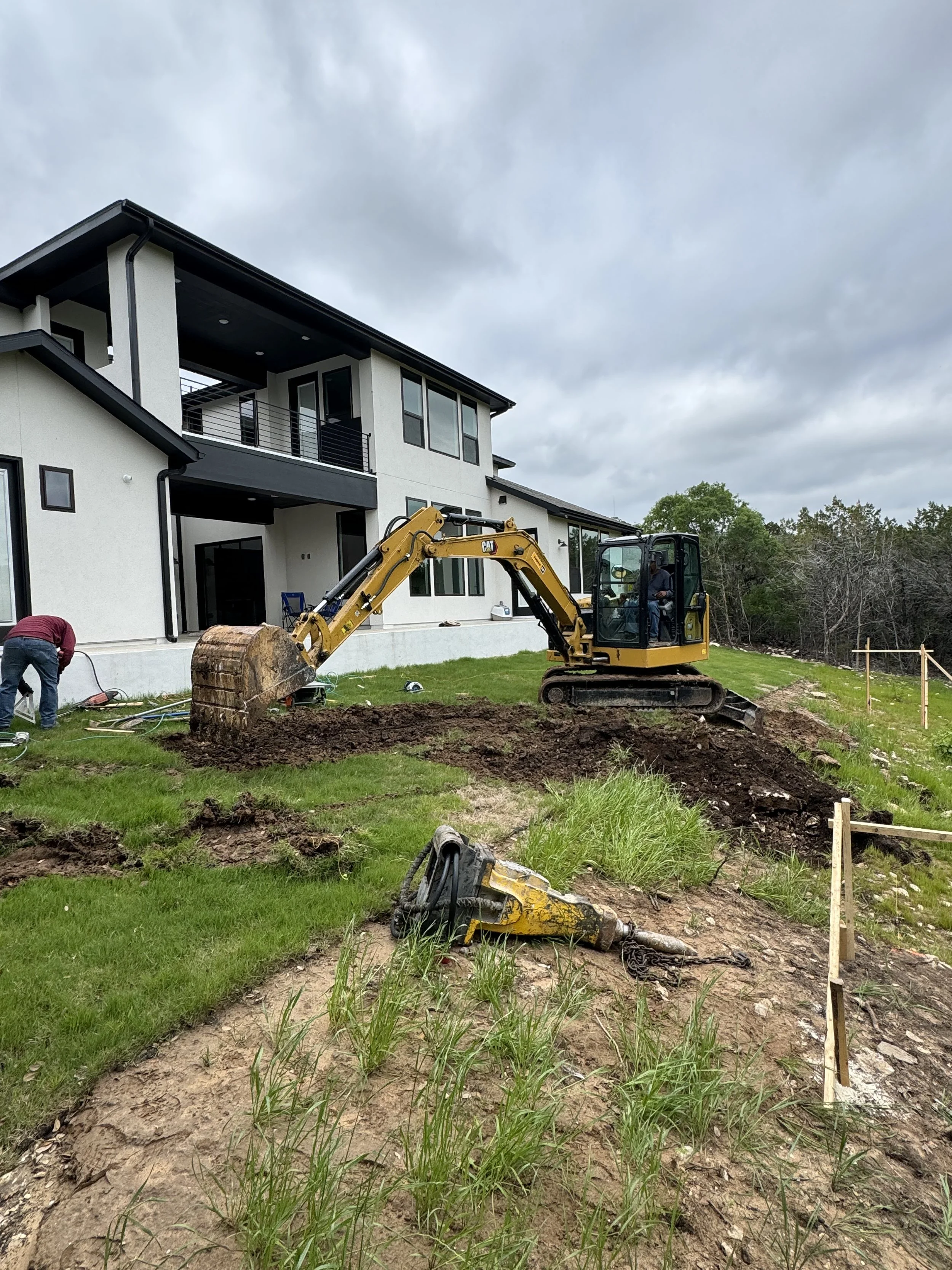 Construction scene with a small excavator digging near a modern two-story house on a cloudy day, with a worker on the left and a removed piece of equipment on the ground.