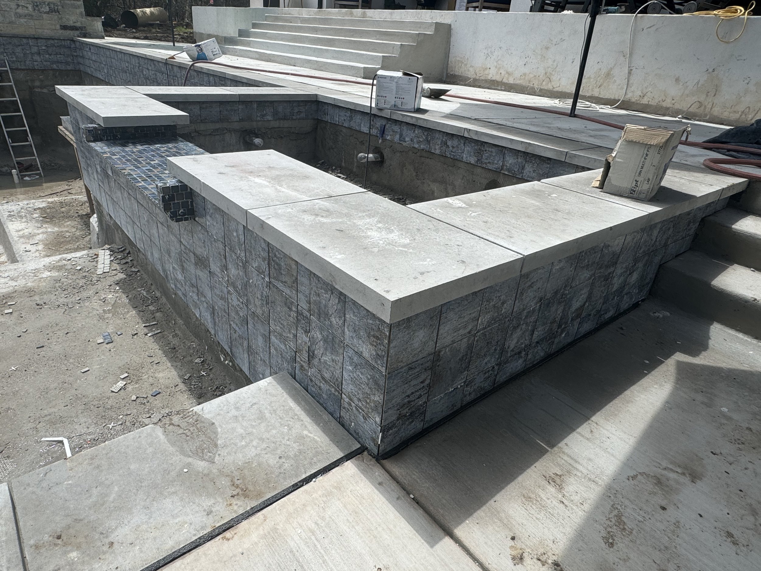 Construction site of an outdoor pool with tiled edges, surrounded by concrete stairs and tools, with some construction materials and equipment visible.