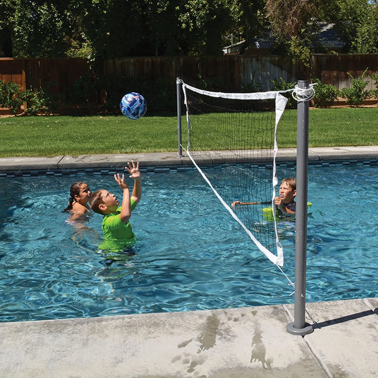 Three children playing water volleyball in a backyard swimming pool with a net, ball, and a wooden fence and trees in the background.