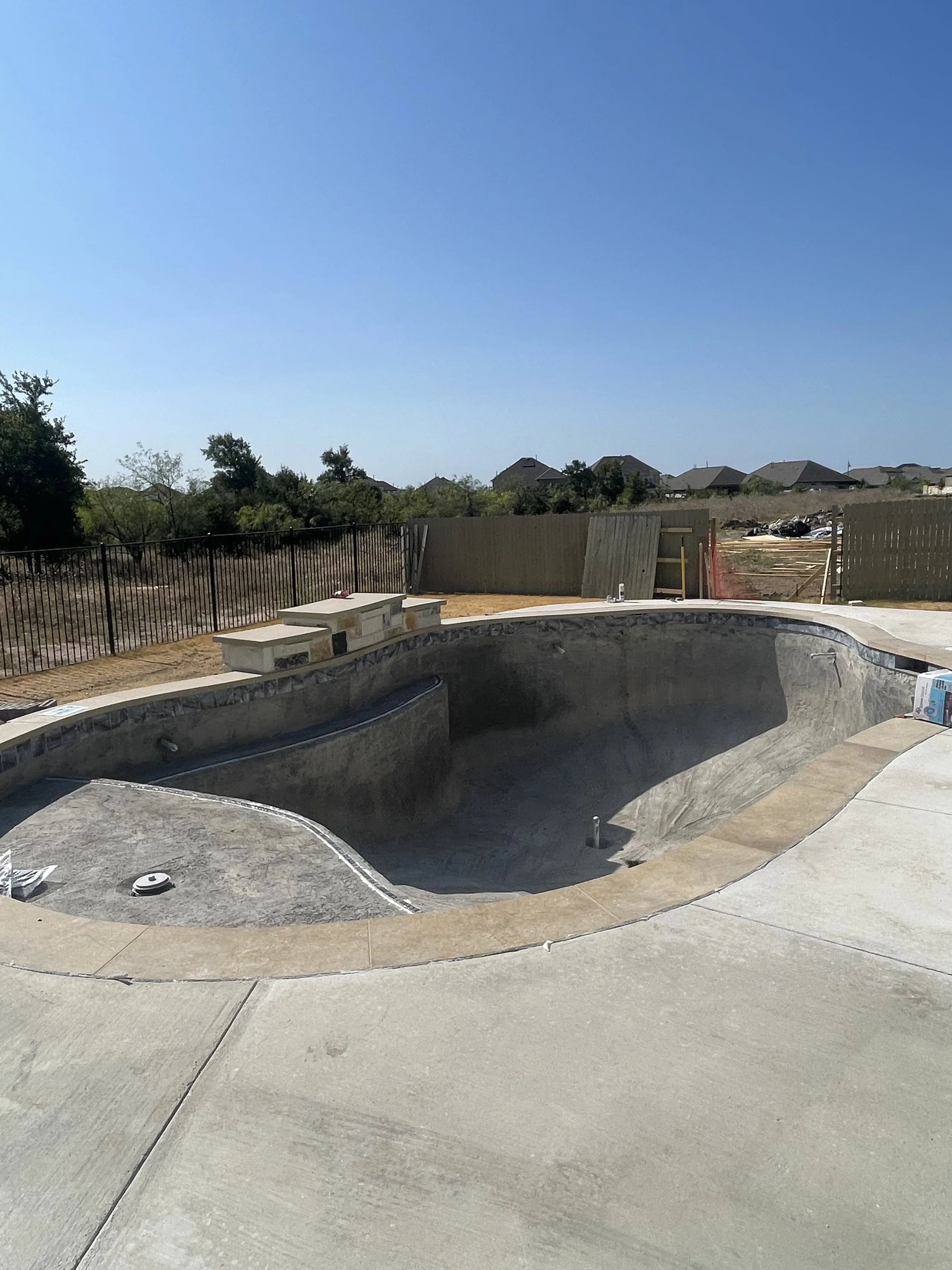Empty backyard swimming pool under construction with concrete surface, surrounded by a concrete deck and fenced yard, in a suburban neighborhood under clear blue sky.